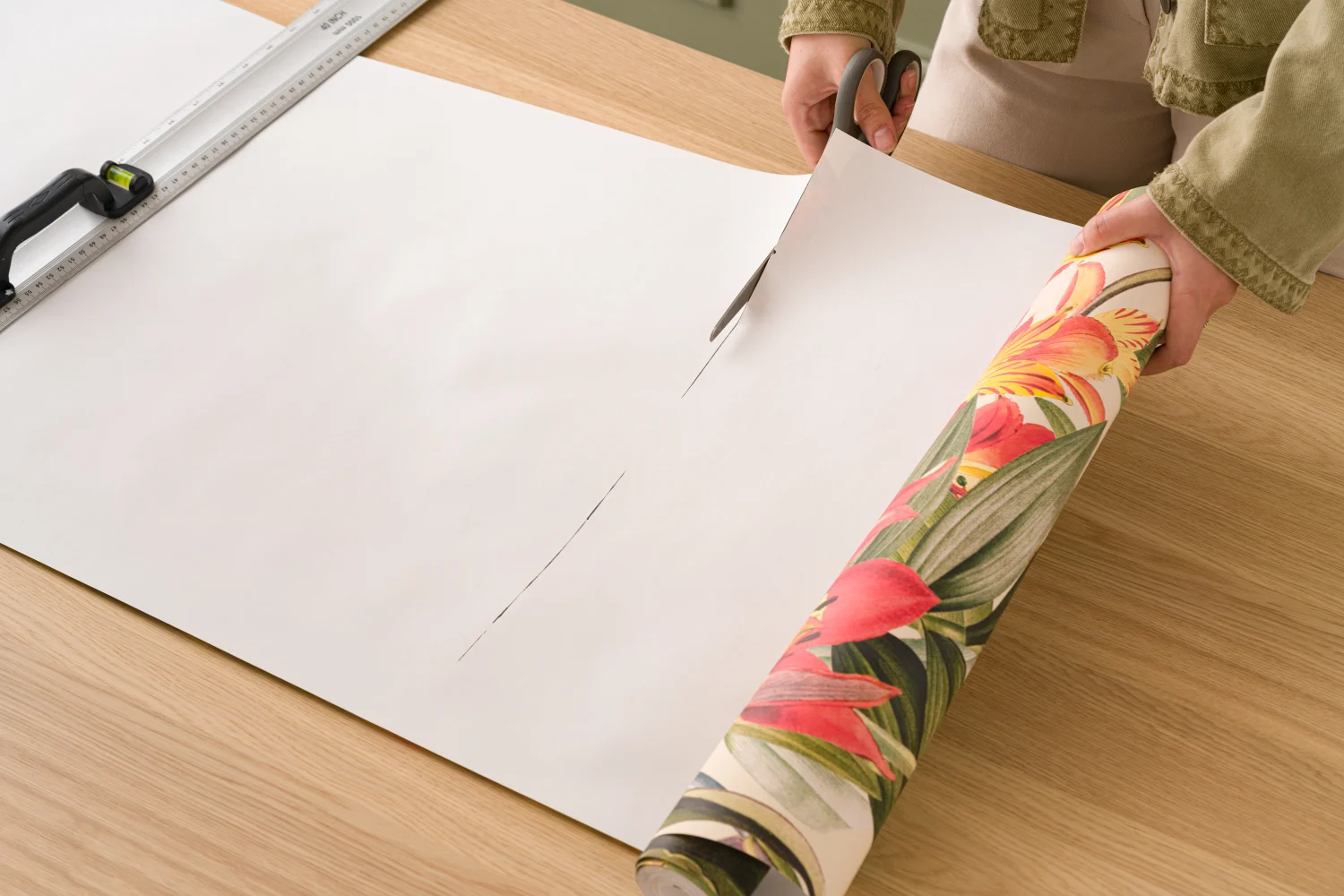 A woman is cutting a roll of vibrant tropical floral wallpaper as part of the process to install the wallpaper.