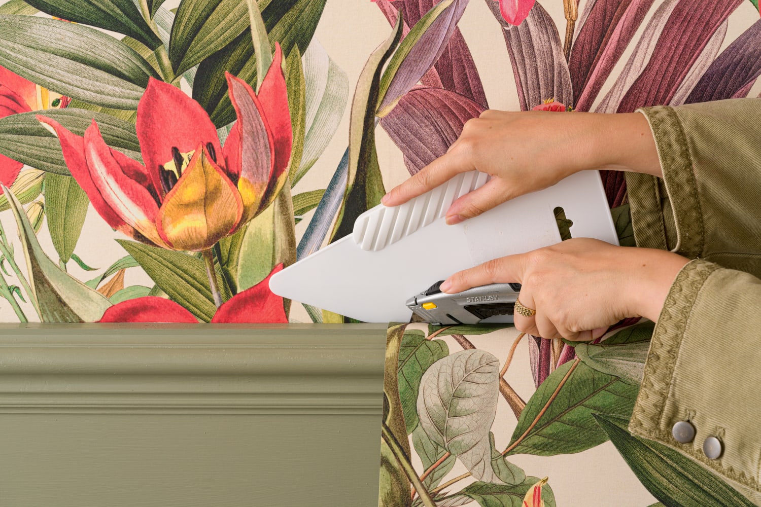 A woman is trimming the excess of a vibrant tropical floral wallpaper as part of the process to install the wallpaper.