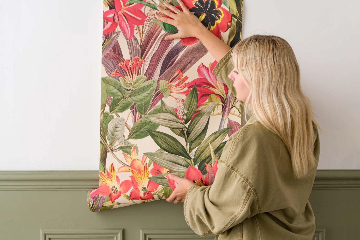 A woman is installing the first panel of a vibrant tropical floral wallpaper with red, yellow, and green hues against a wall with sage green paneling.