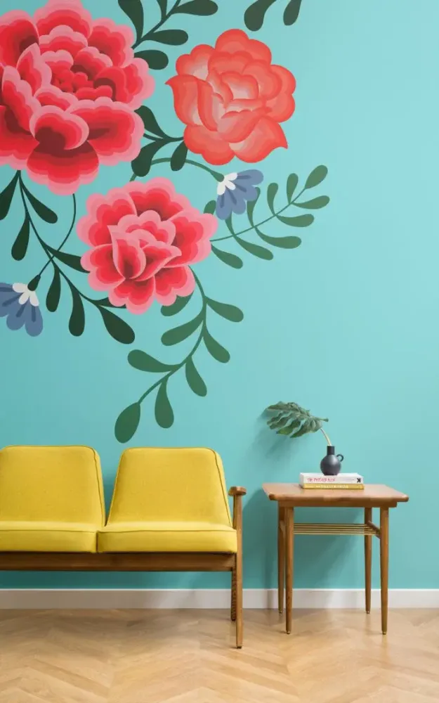 A stylish interior featuring a mustard yellow couch and a small wooden side table with a vase and books, set against a pastel teal wall adorned with large pink floral designs.