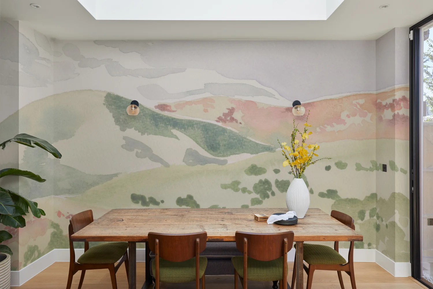 A hand-painted mural of abstract hills and valleys in earthy pastels decorates a dining space with wooden furniture and greenery.