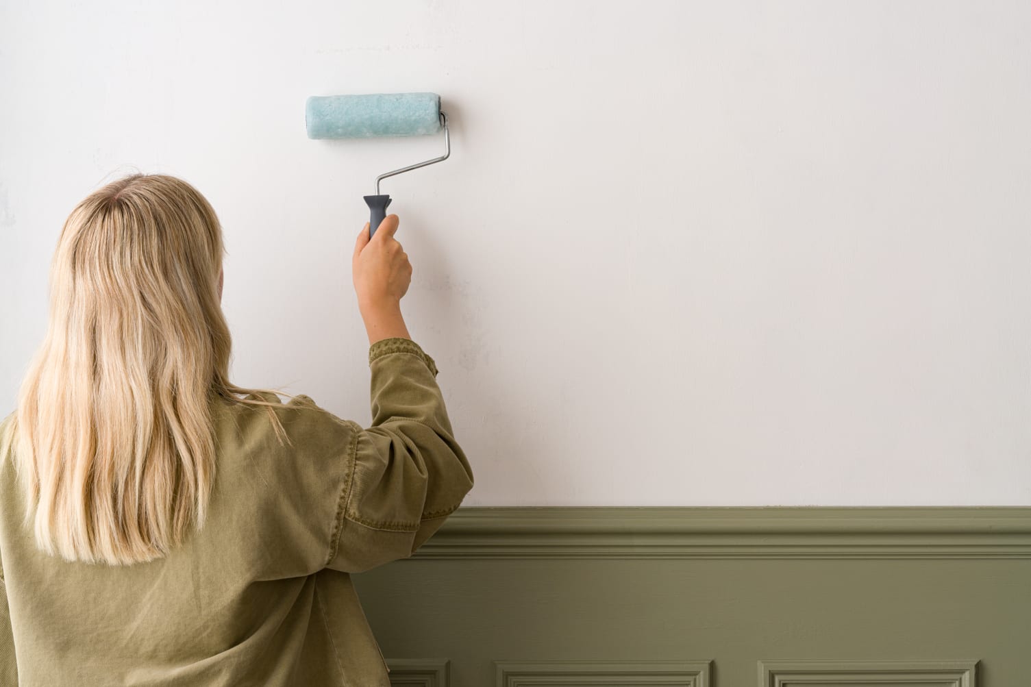 A woman is applying wallpaper paste to a wall with a paint roller as part of the process to install the wallpaper.