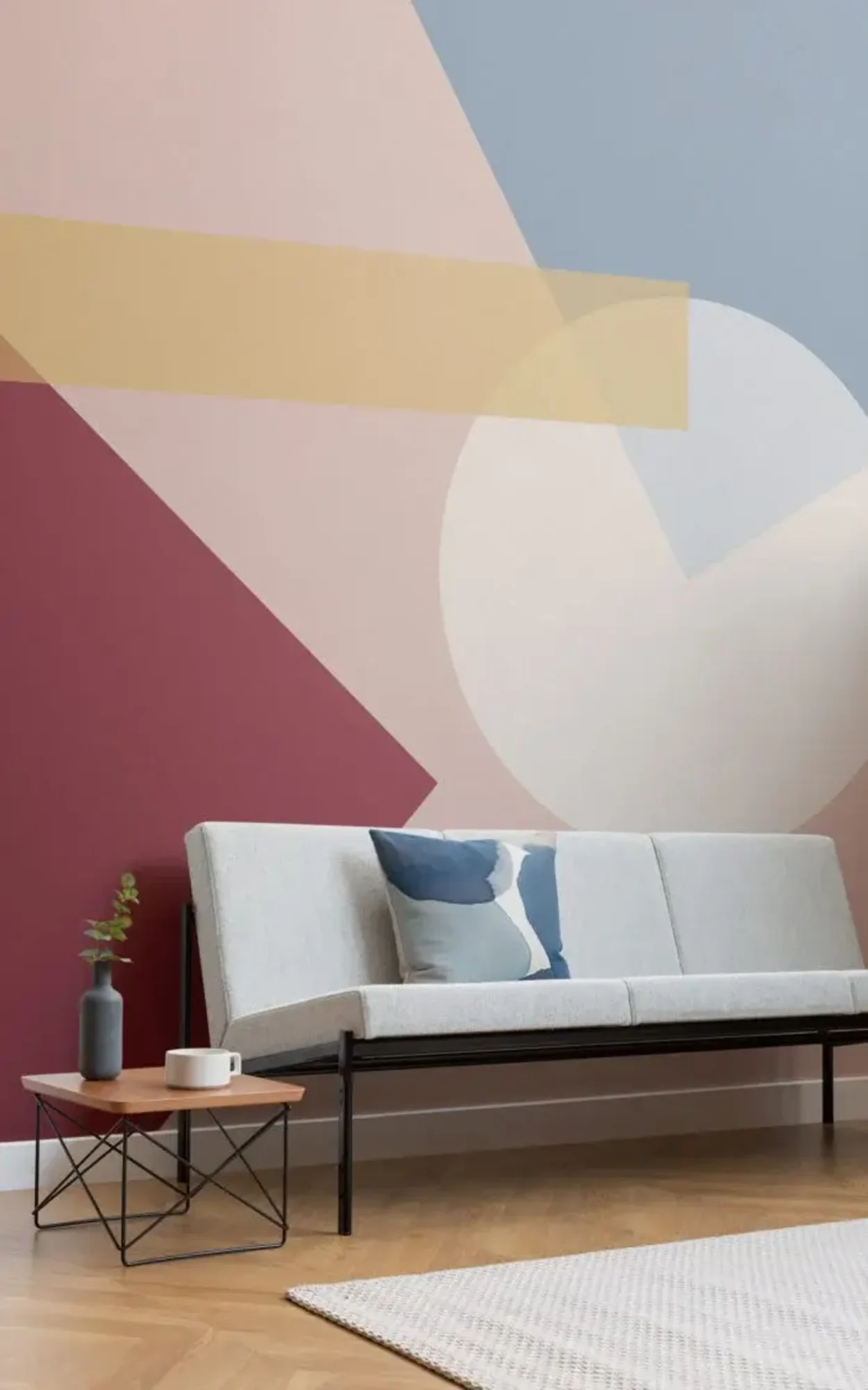 A modern living room with a light gray sofa against a vibrant geometric patterned wall in shades of pink, red, and yellow, complemented by a small wooden table with a decorative vase and a rug.