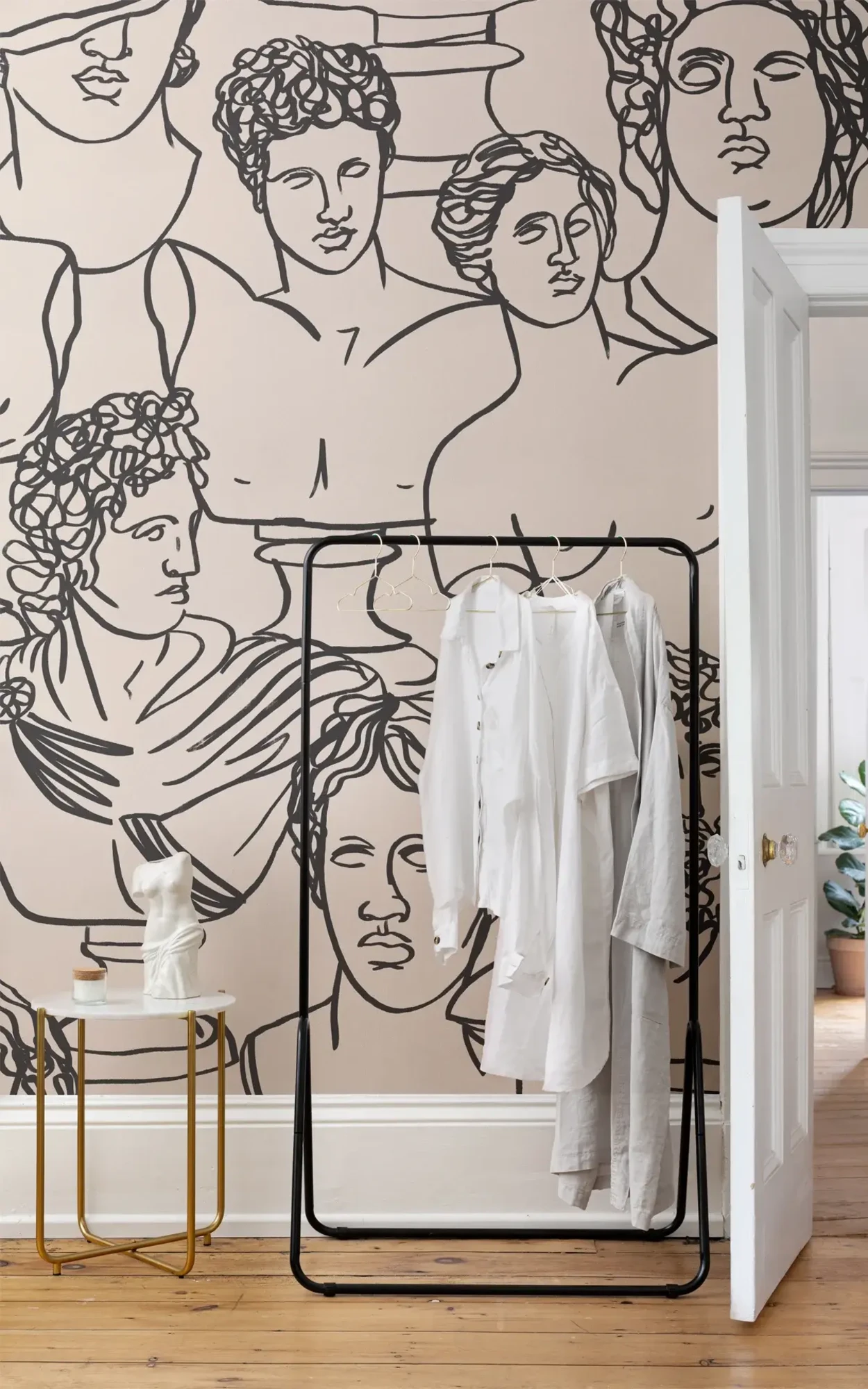 A stylish room corner featuring a unique mural of classical Greco-Roman busts in black line art on a beige wall, complemented by a simple black clothing rack with white shirts and a small gold side table.