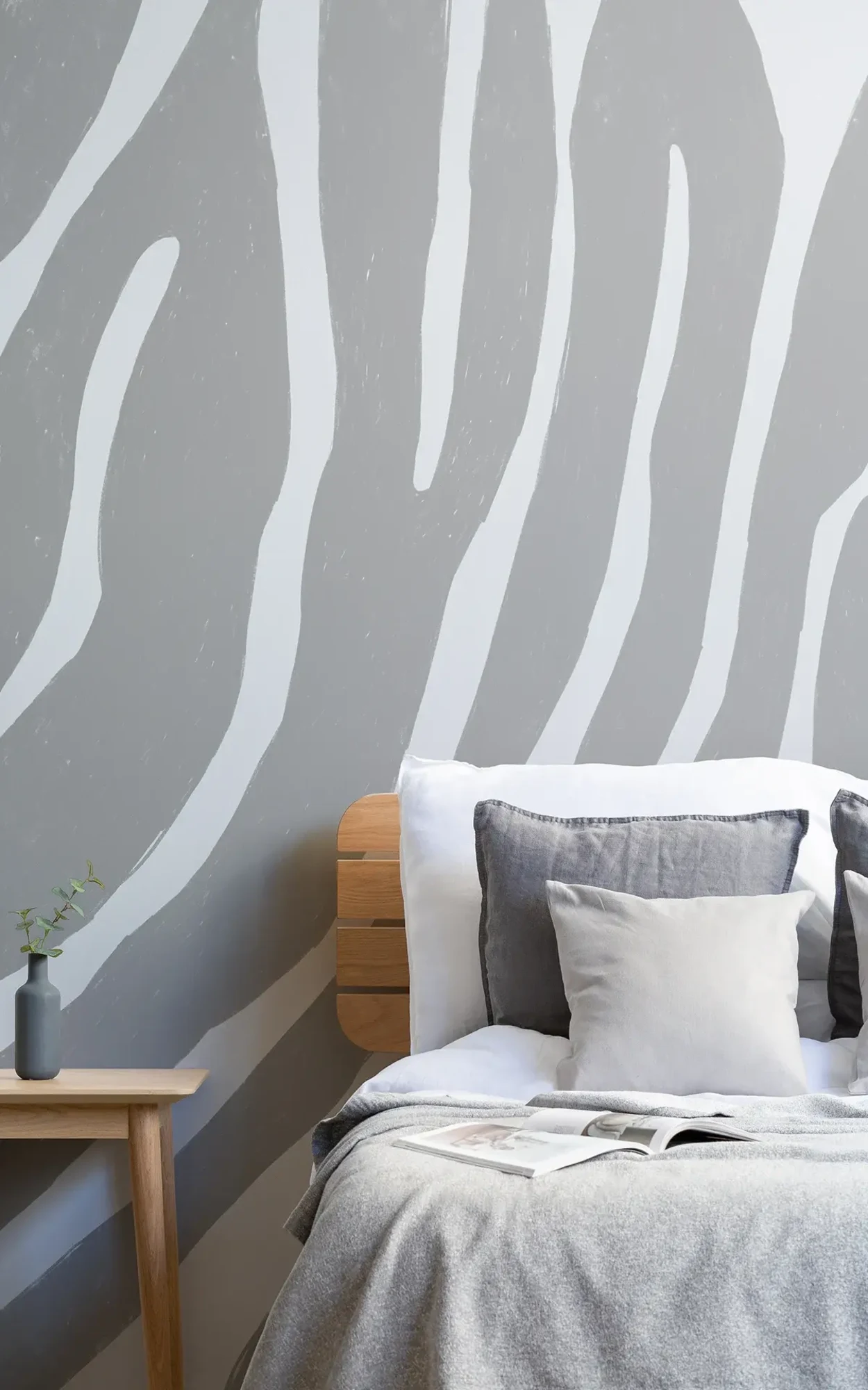 A modern bedroom features a wooden bed with gray and white bedding, a small wooden side table with a vase, and a wall with a large gray zebra stripe pattern.