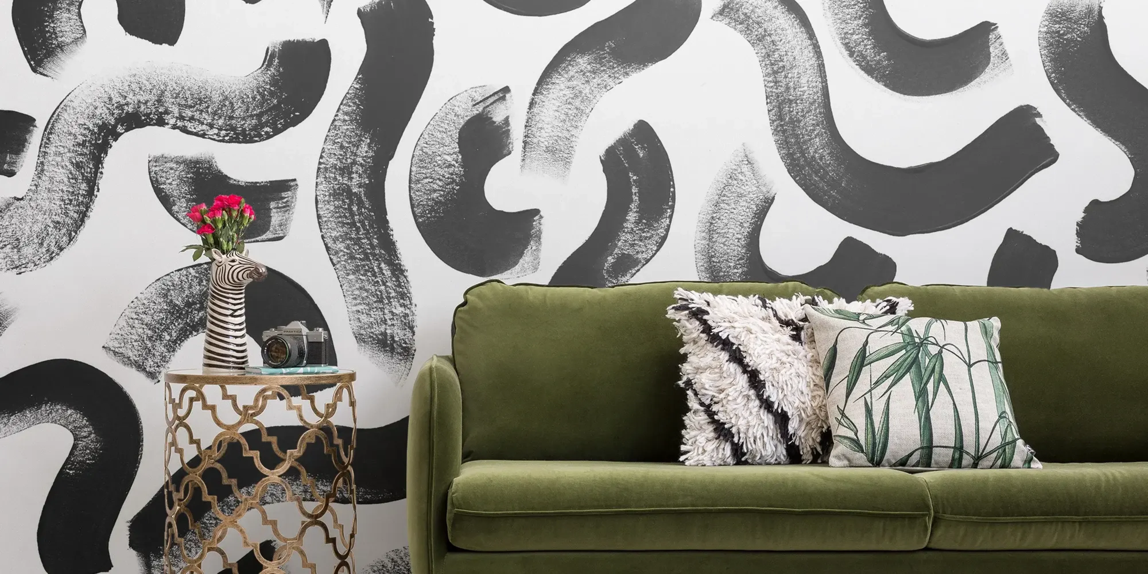 A stylish living room features a green velvet sofa with patterned cushions, a small ornate side table holding a zebra-shaped vase with pink flowers, and a black-and-white abstract wall mural in the background.