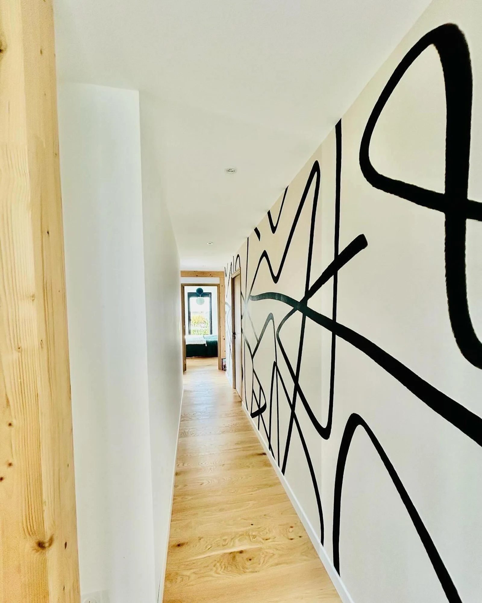 A modern hallway with a bold black abstract line mural on a white wall, light wood flooring, and a door leading to a bright room.