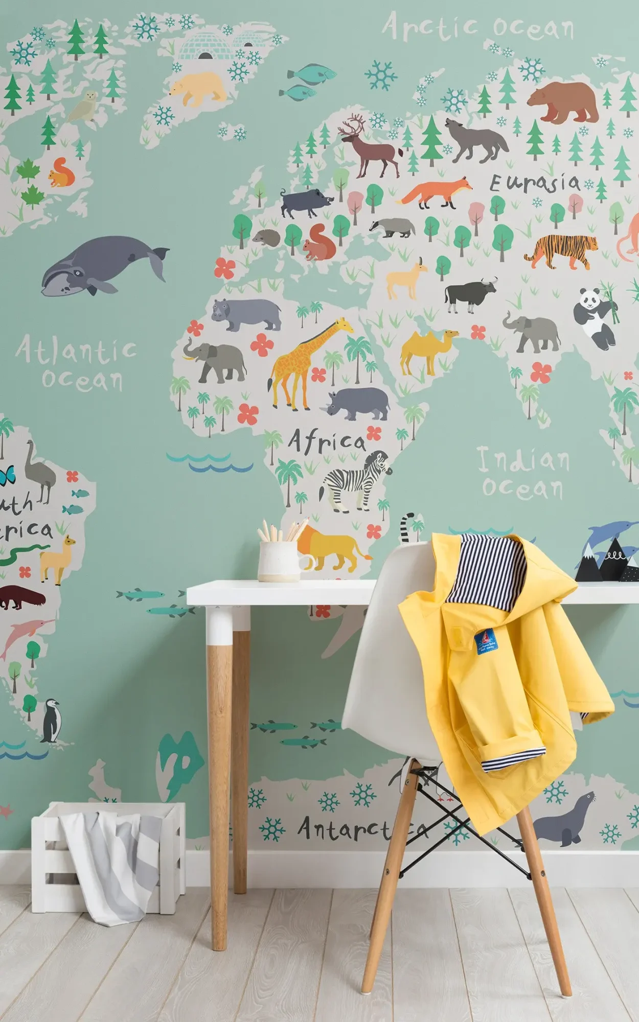 A colorful children's room corner featuring a world map wallpaper with illustrations of various animals and regions, a white desk with a chair and a hanging yellow jacket, and wooden flooring.