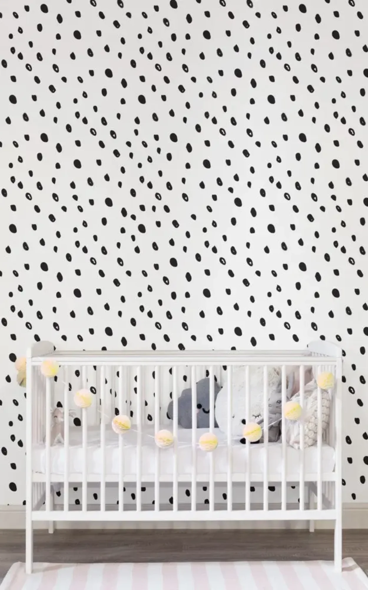  A nursery features a white crib with stuffed animals and a string of yellow pom-poms, set against a black-and-white speckled wall and a pink striped rug.