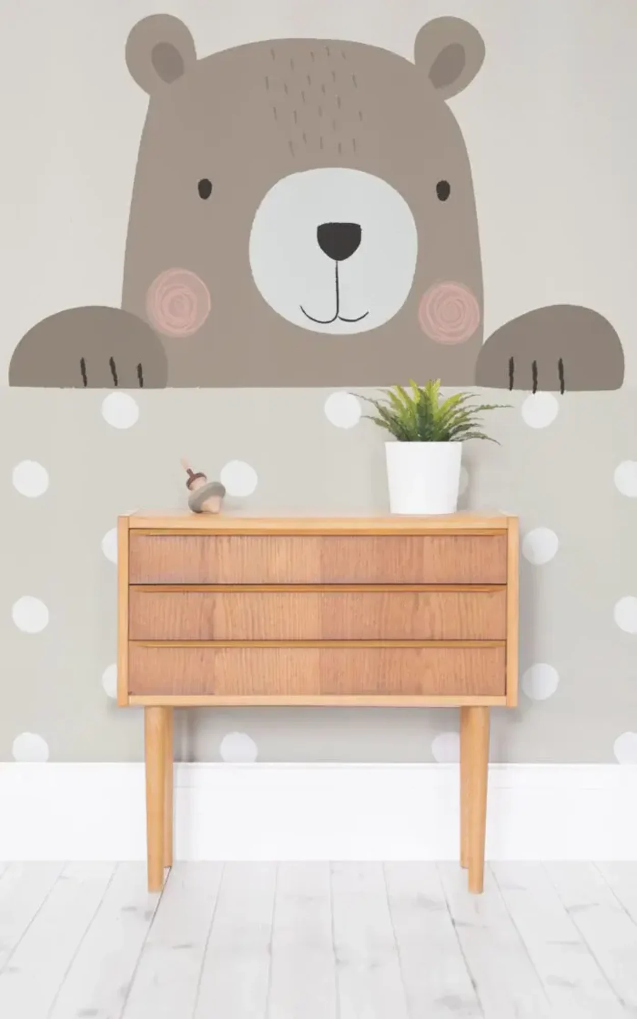 A playful children's room features a wooden dresser, a potted plant, a small toy, and a large mural of a cute bear peeking over polka-dotted wallpaper.