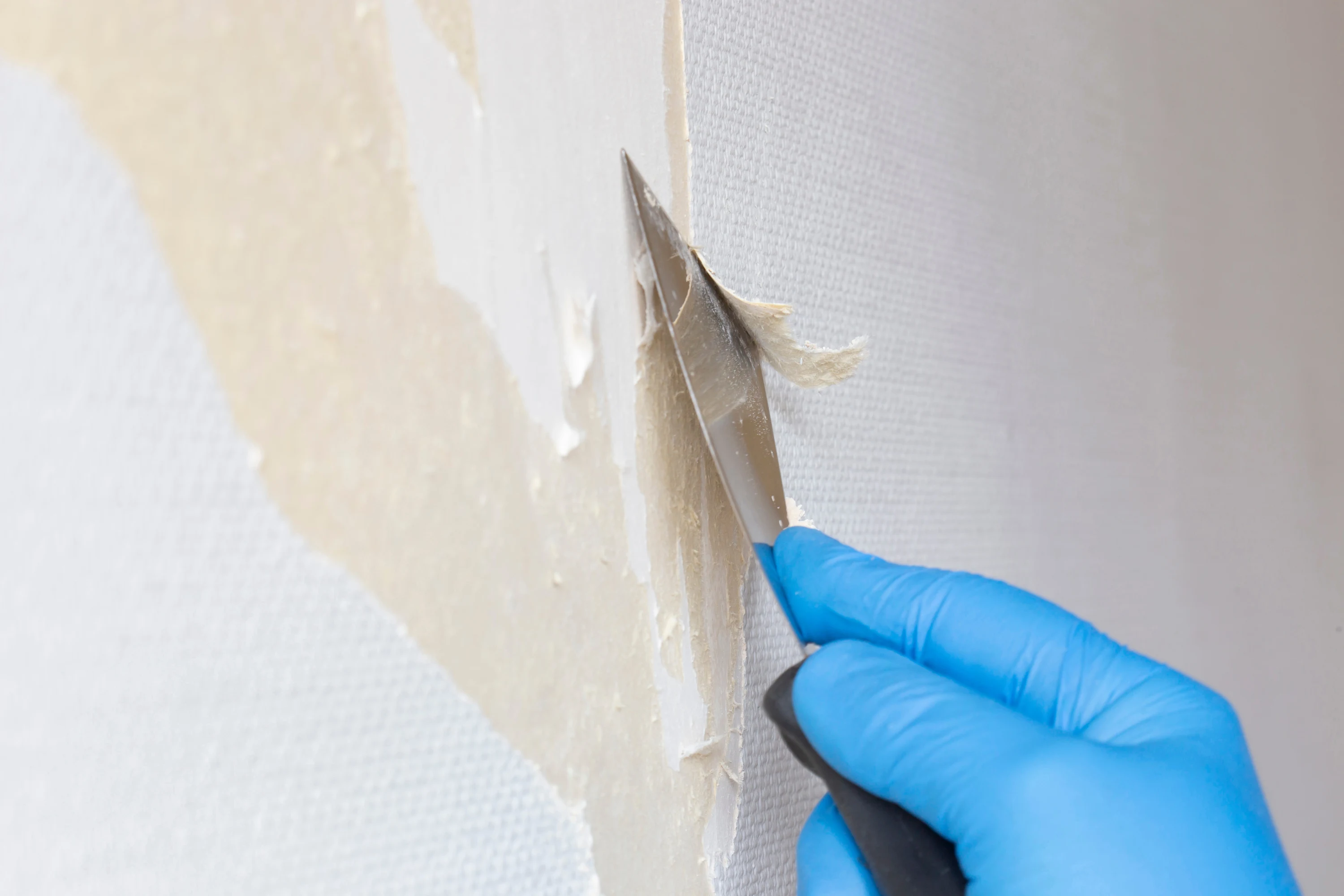 Gloved hand using a scraper to remove old wallpaper from a wall.