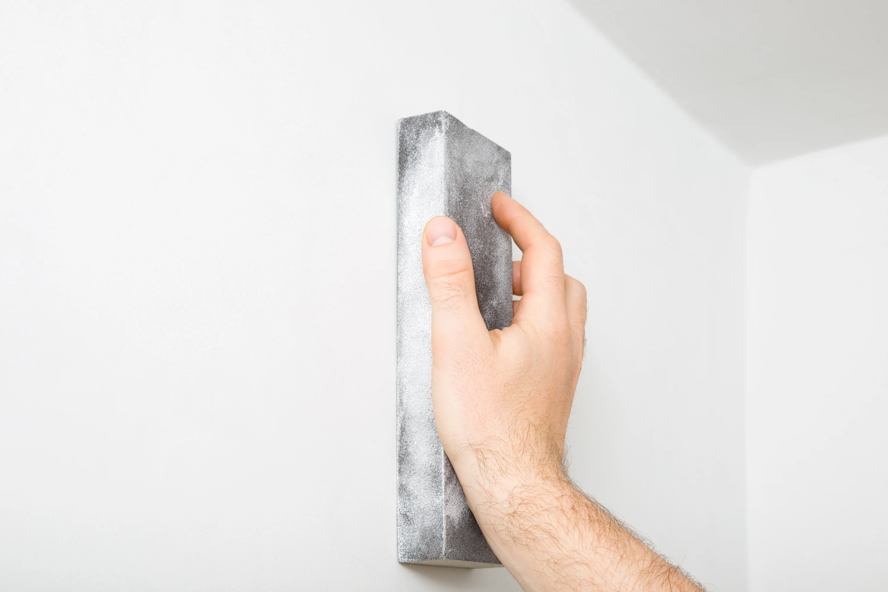 Hand holding a sanding block against a white wall for surface preparation.