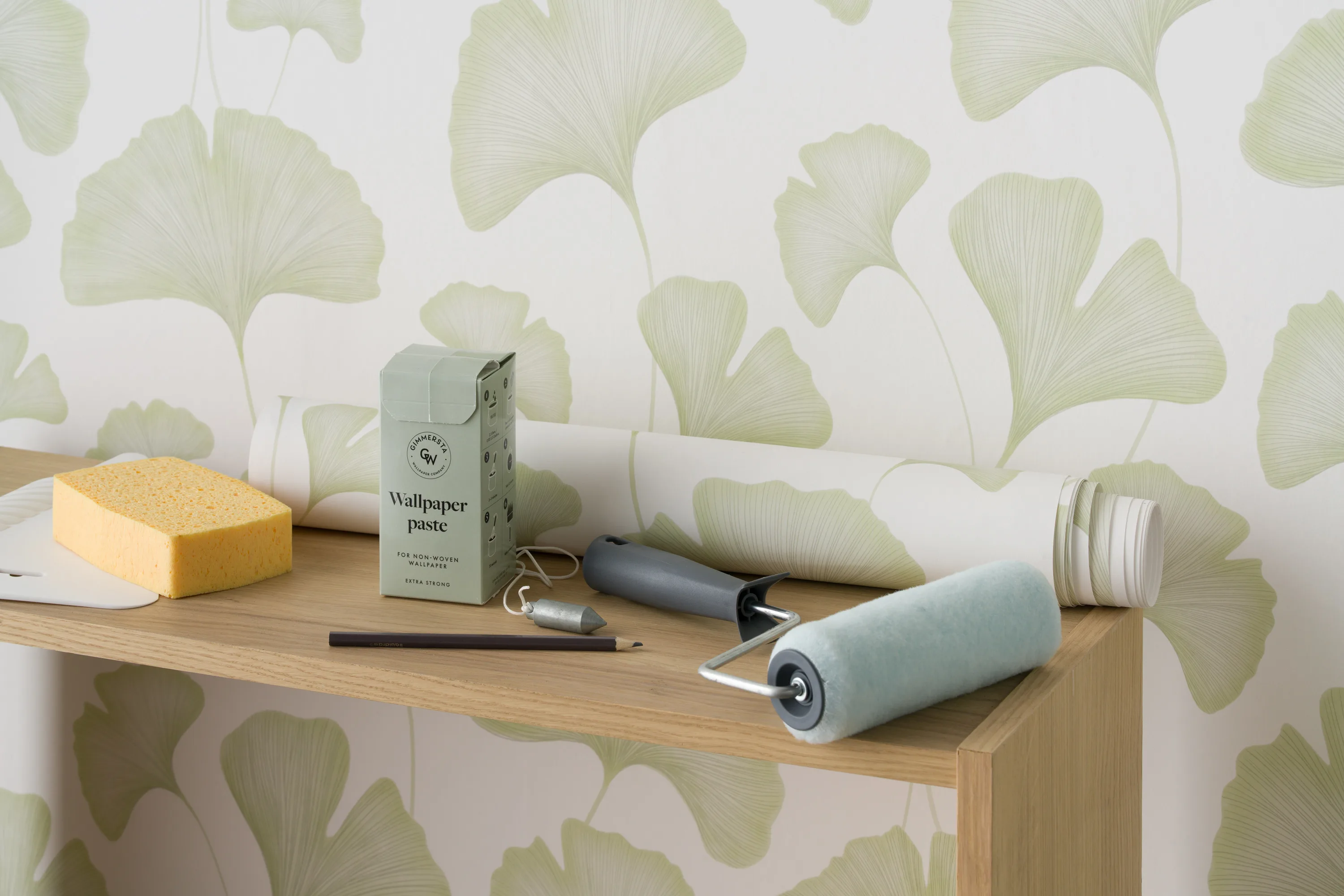 Wallpaper tools and paste box arranged on a wooden shelf against a leafy green wallpaper.