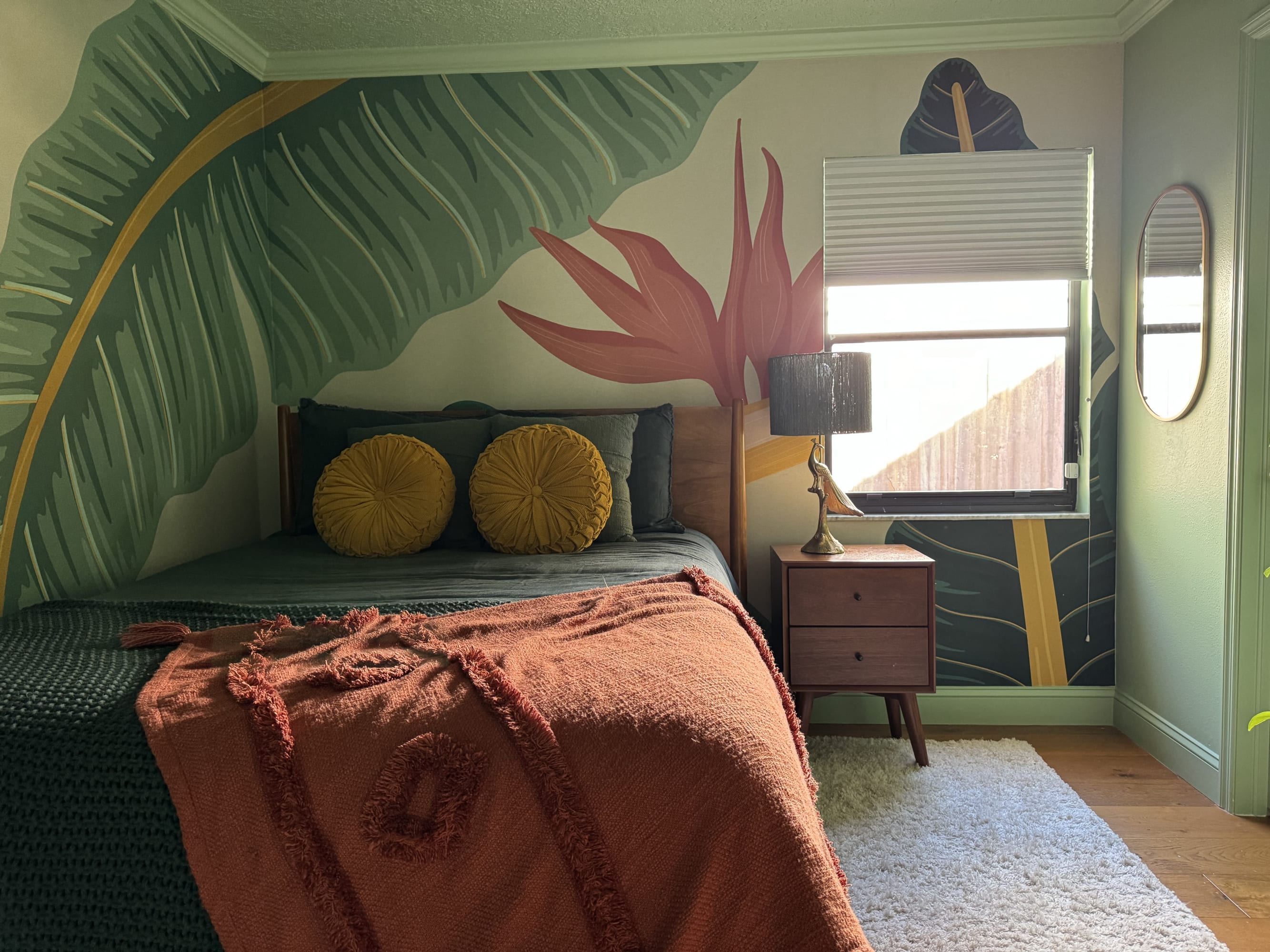 A bedroom featuring a tropical-themed mural with large leaves and flowers, a green bed with yellow pillows and terracotta blanket, a window with the blinds open and a wooden nightstand with a bird shape lamp.