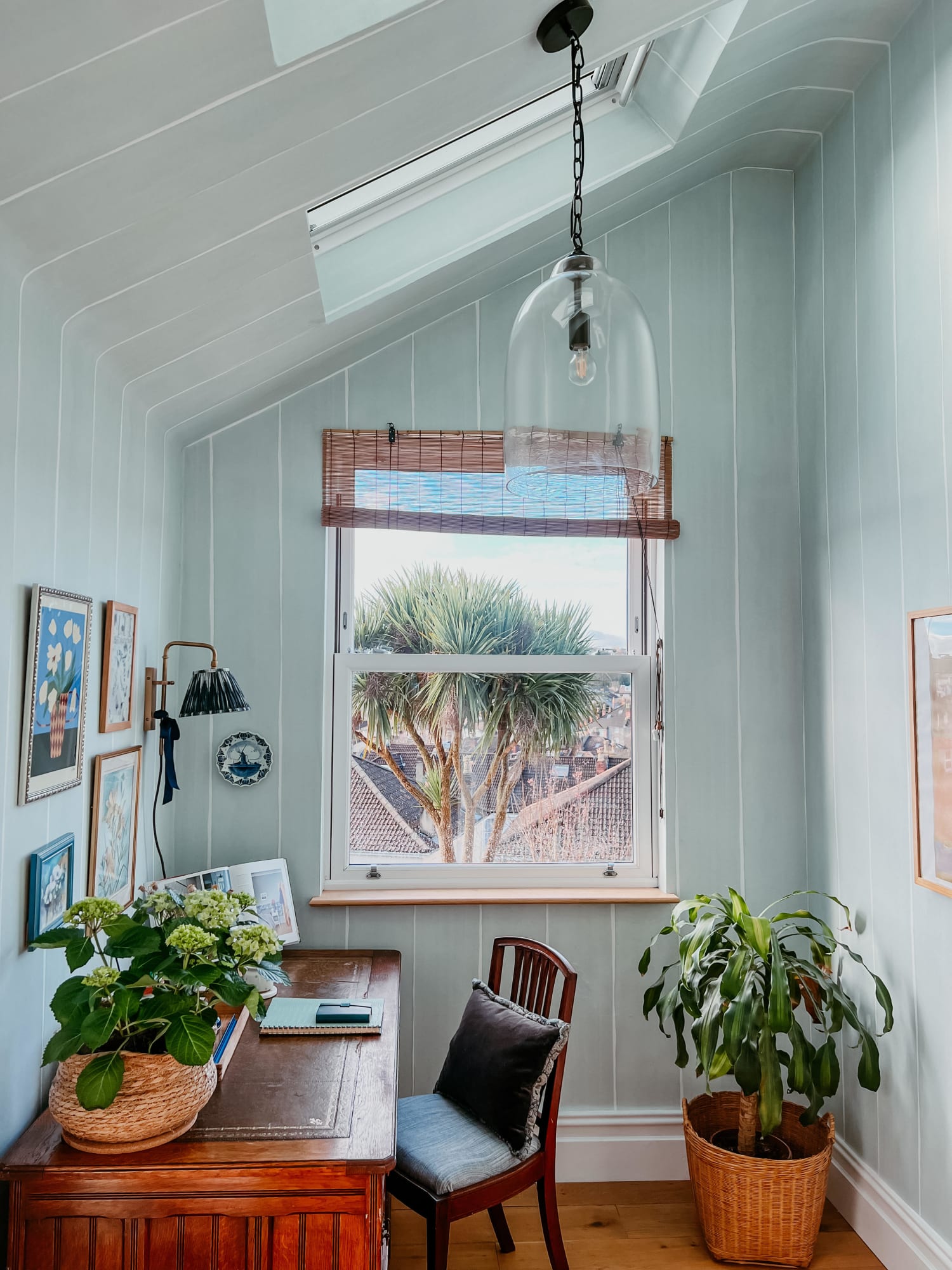 The image shows a small, cozy workspace with pastel striped wallpaper and a window overlooking a palm tree, featuring a wooden desk, plants, and framed art.