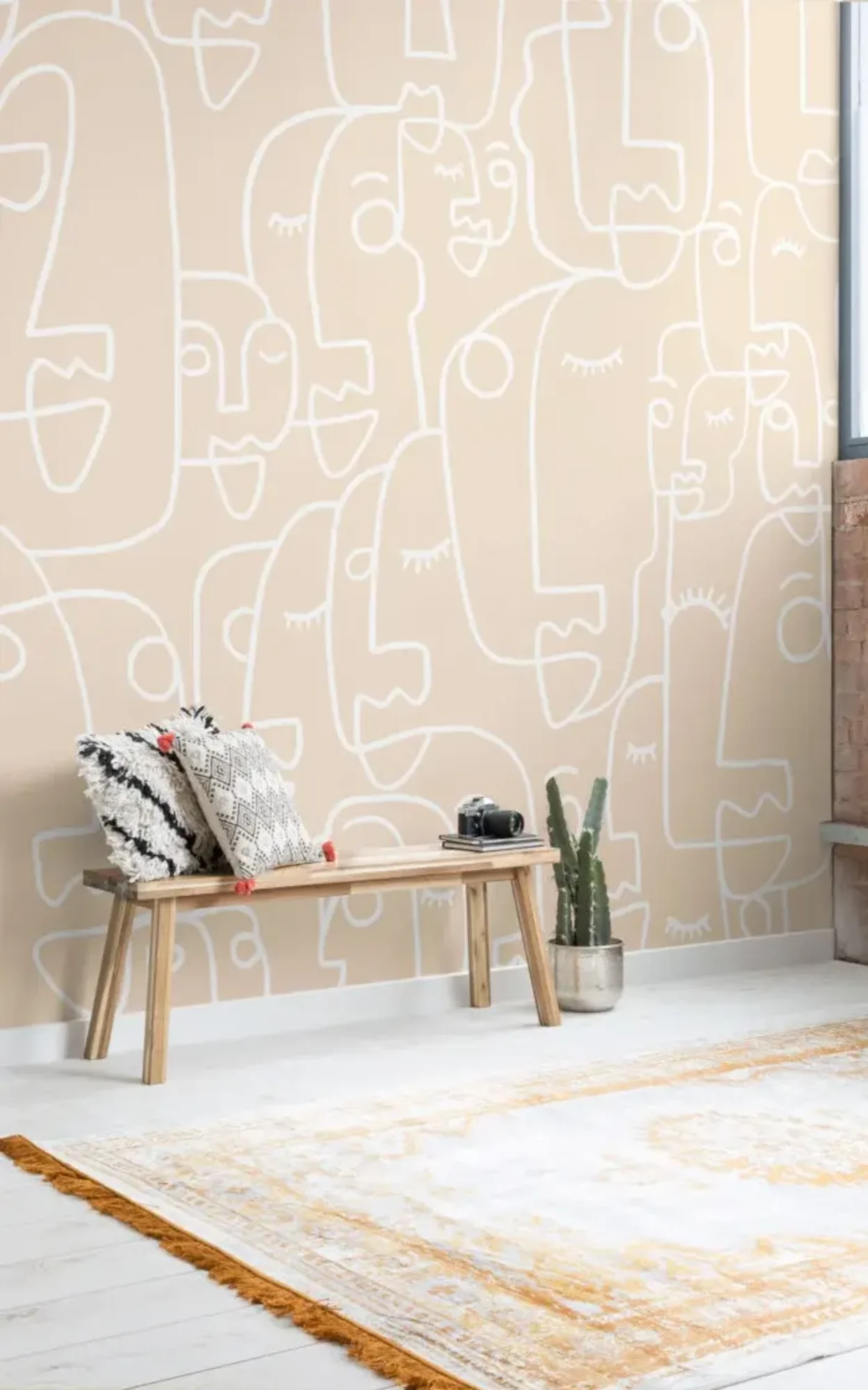  A cozy room features a wooden bench with decorative pillows, a potted cactus, and a beige wall with white face doodles.