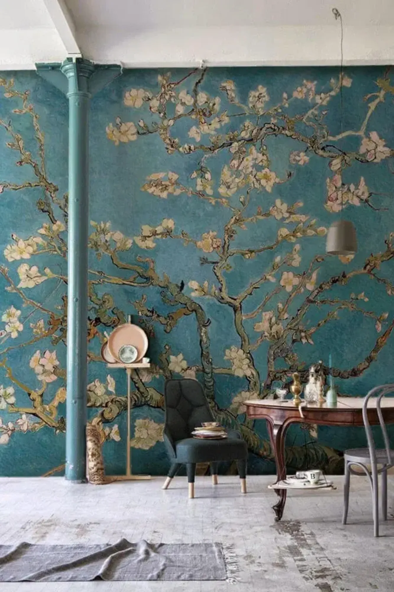 A vintage-style room with teal floral wallpaper, featuring a dark green armchair, an ornate wooden desk with vintage items, a metal chair, and a large teal structural column, all set on a distressed gray rug.