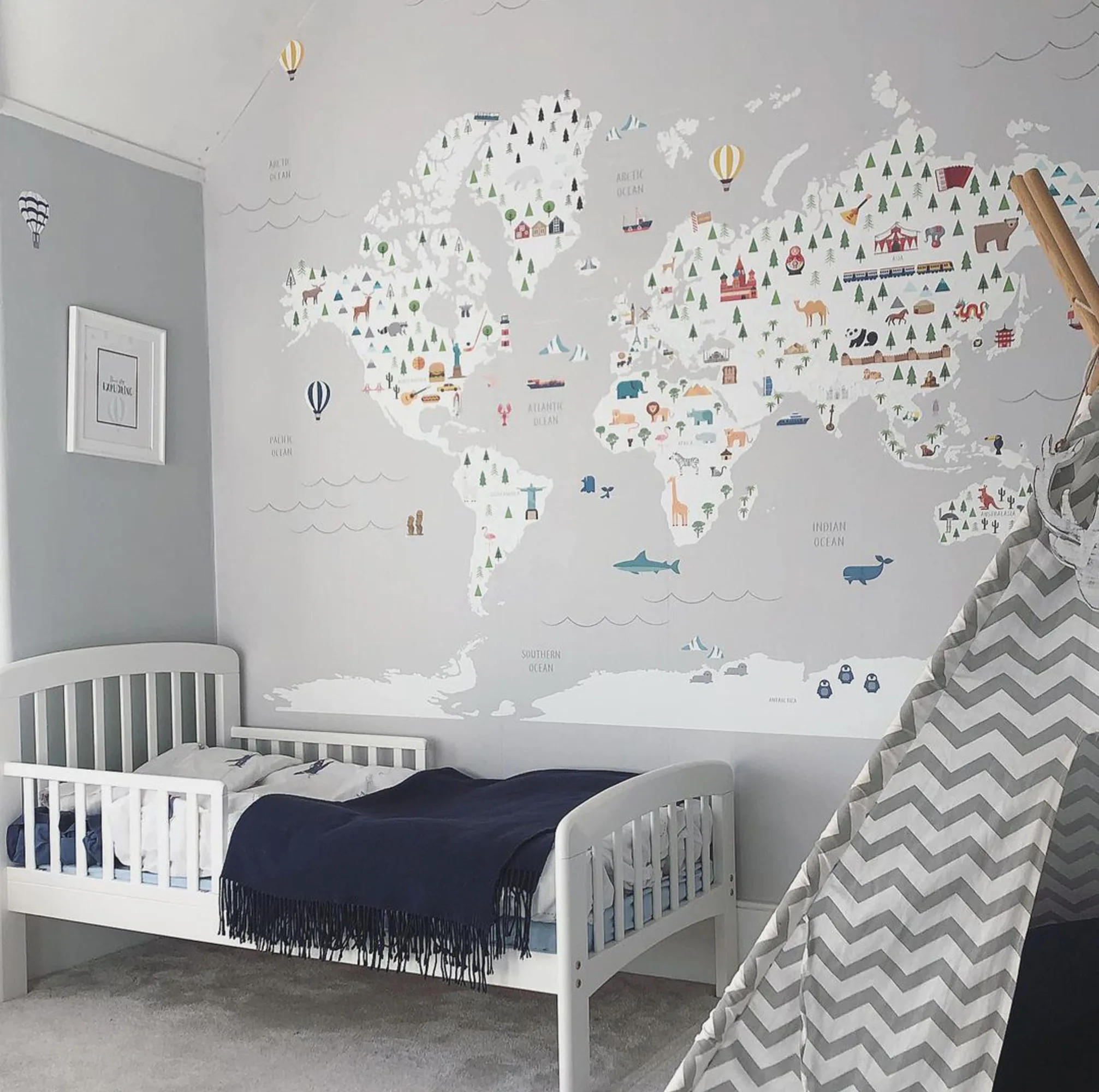 A world map mural in a gray and white color scheme, decorating a child's bedroom with a white bed and a chevron-patterned teepee.