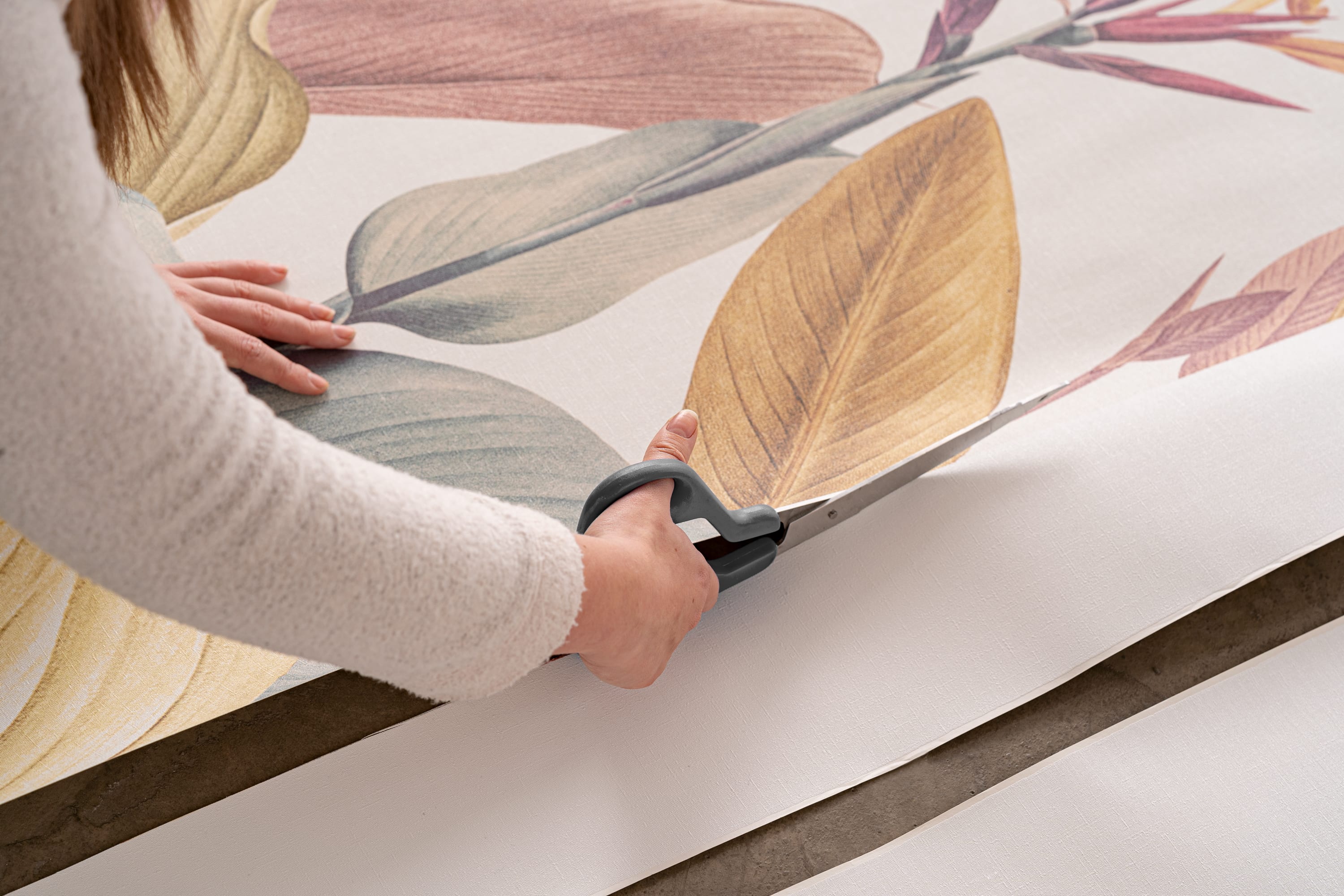 A person uses scissors to cut a panel of tropical botanical wallpaper on a table.