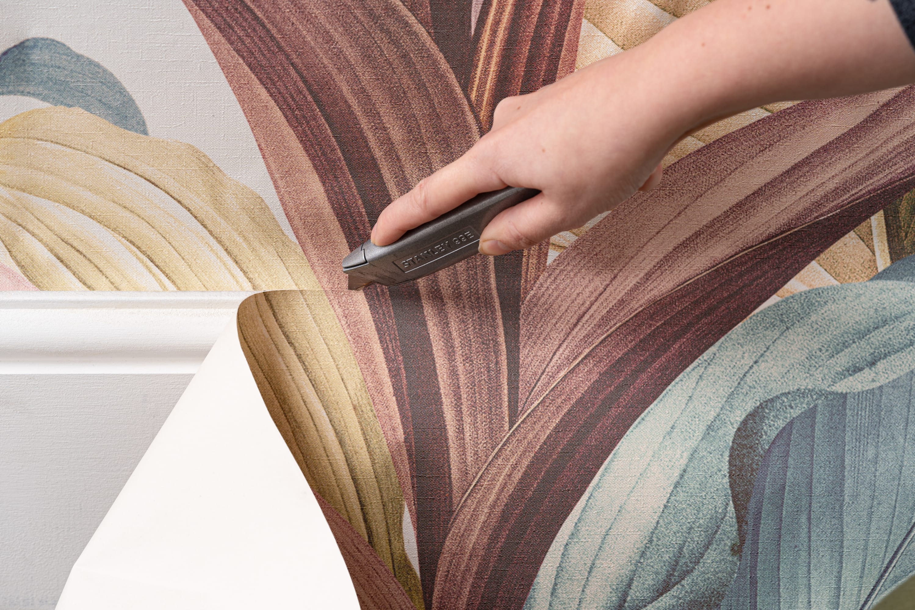 A hand uses a utility knife to trim colorful botanical wallpaper along the wall edge.