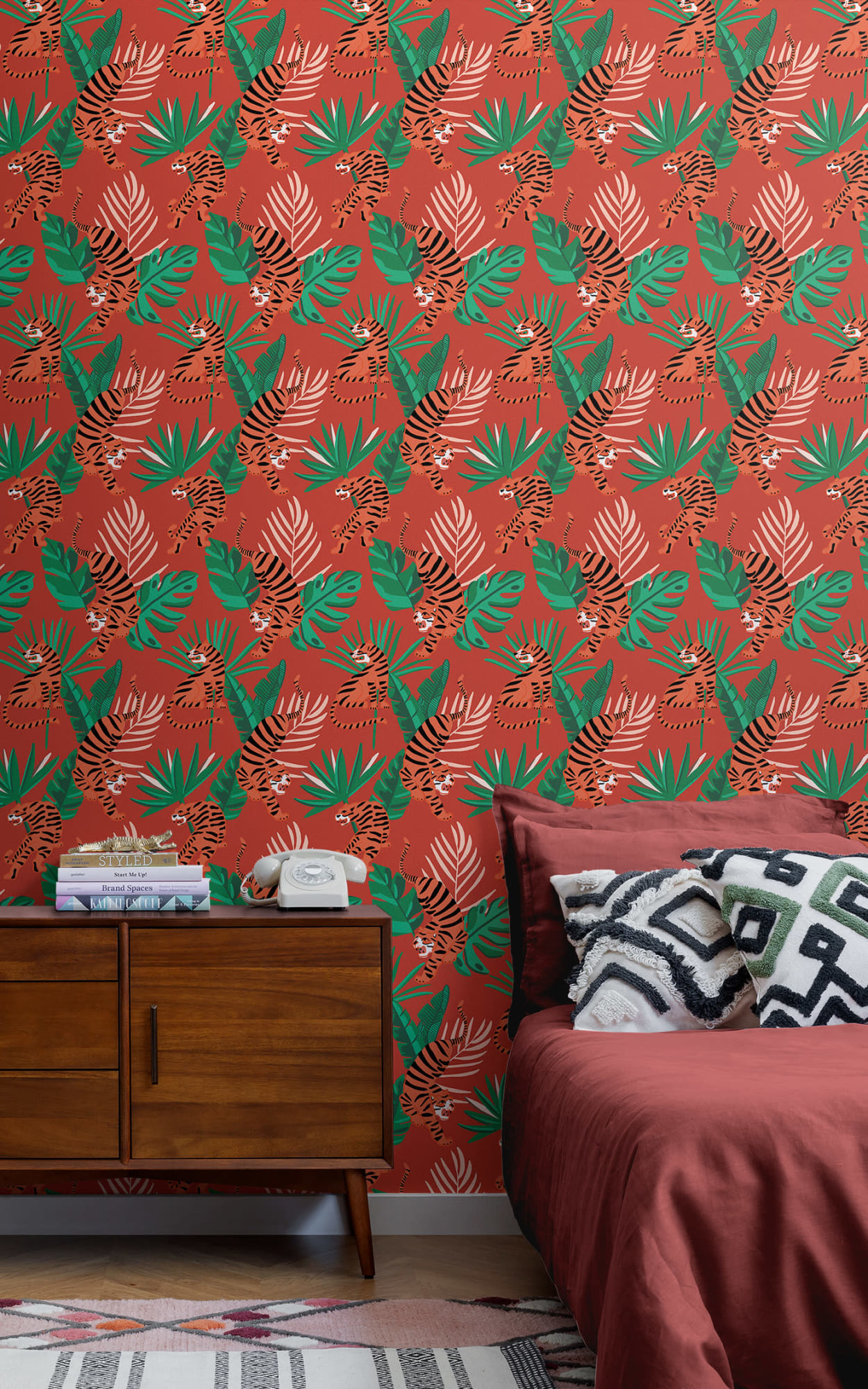 A vibrant bedroom scene with a bold red and green tropical leaf and tiger patterned wallpaper, featuring a dark wooden bedside table, a white vintage telephone, and a cozy bed with a deep red duvet and decorative pillows, set over a patterned pink rug.