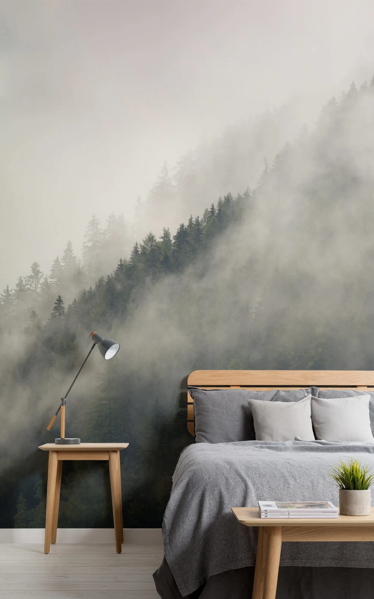 Bedroom with a wall mural of a foggy pine forest. Features a wooden bed with gray bedding, a side table with a lamp and a small plant, set on a light wooden floor.