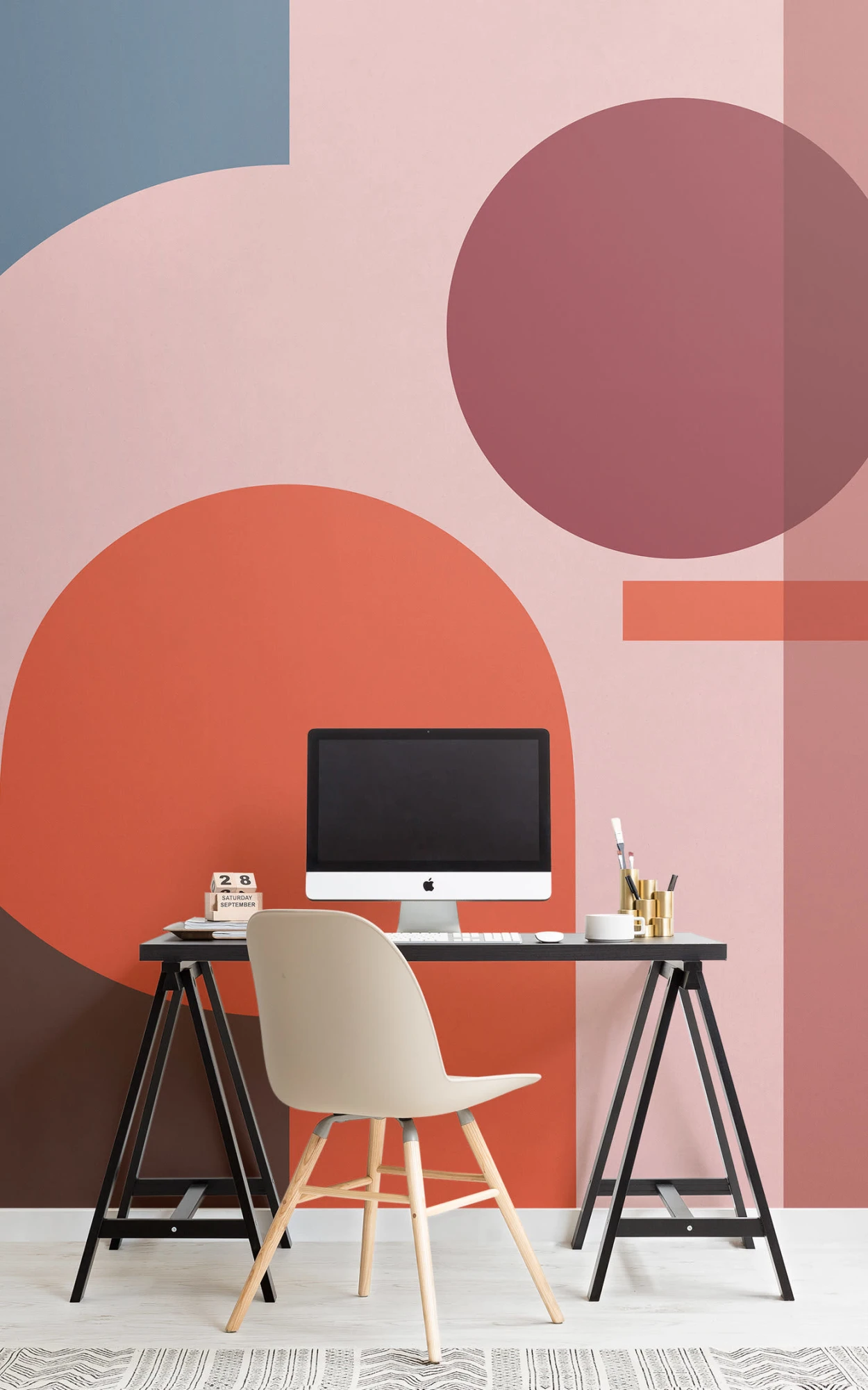 Modern workspace with a geometric wall design in shades of pink and red. Features a simple desk with a computer, a beige chair with wooden legs, and small desk accessories like pens and a calendar.