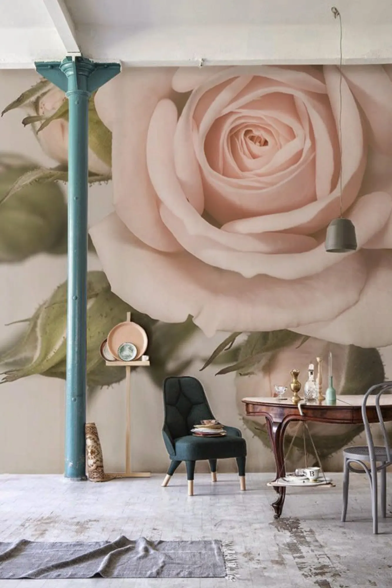 Eclectic room with a large wall mural of a pale pink rose. Features a teal armchair, a vintage wooden desk with assorted items, and a small metal chair. A teal column and white floor accentuate the space.