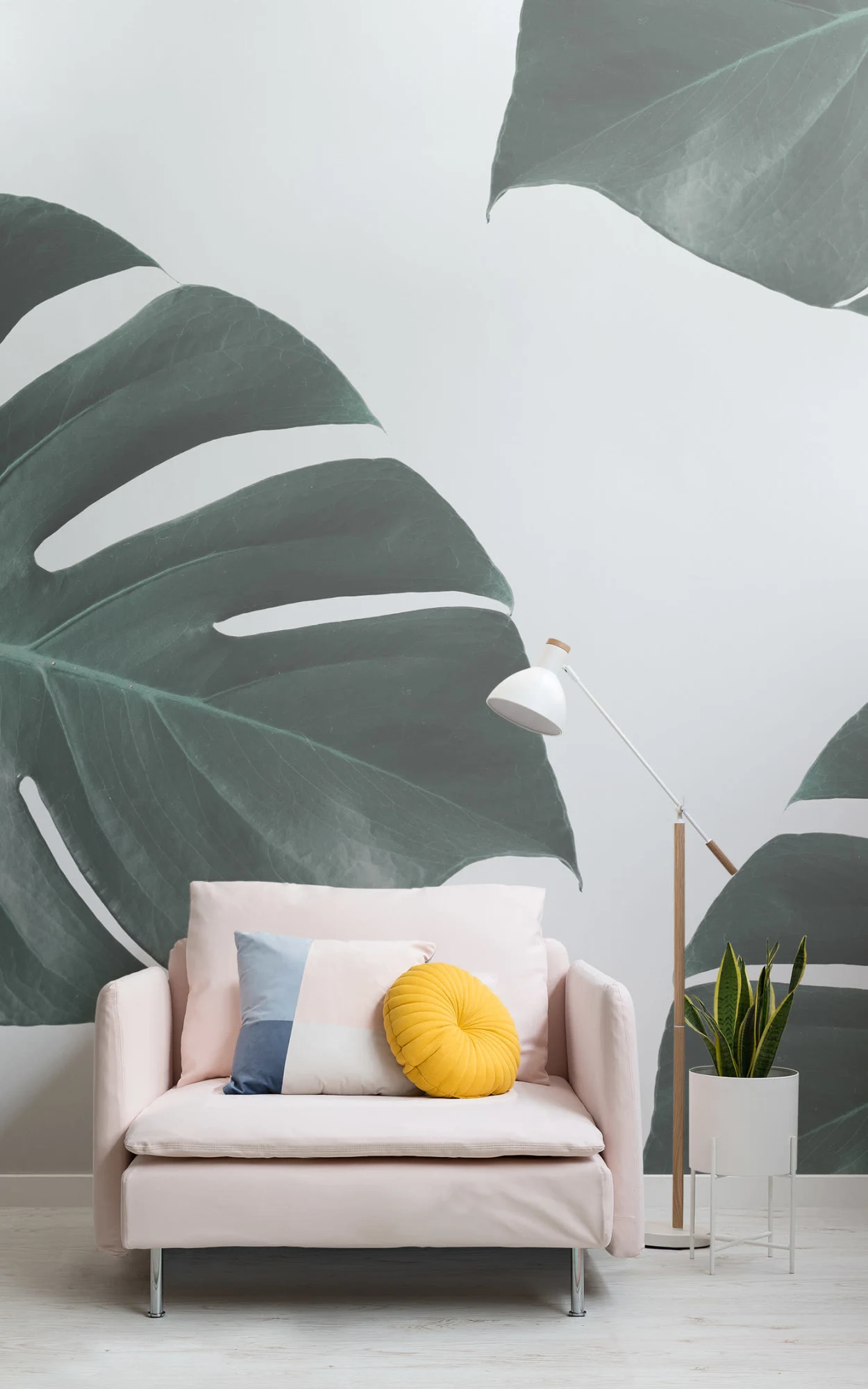 Modern living room with a large wall mural of monstera leaves. A pale pink sofa with a multicolored pillow and a yellow round cushion is paired with a white floor lamp and a potted snake plant.