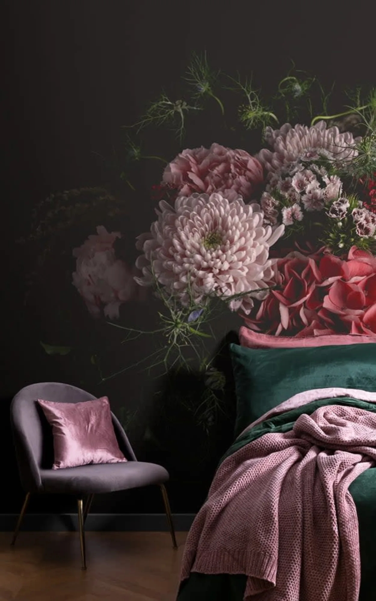 A moody bedroom corner featuring a large floral mural with lush pink and red flowers on a dark background. A velvet chair with a soft pink cushion sits beside a bed with dark green sheets and a knitted pink throw blanket.