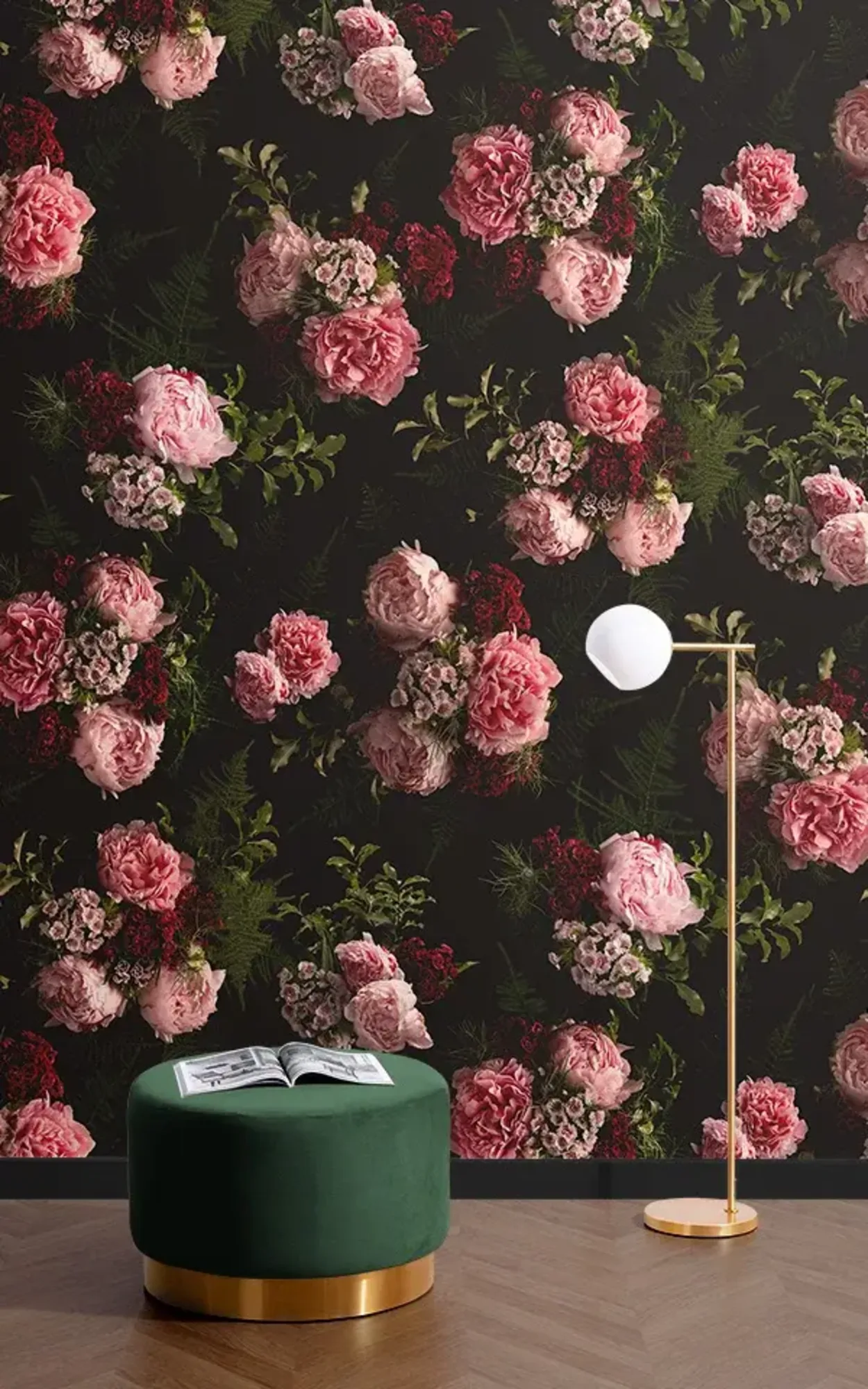 A reading nook with a wall adorned by a detailed floral wallpaper featuring large pink and red roses on a dark background. A dark green velvet ottoman with a gold base is centered in the space, topped with an open book, next to a gold floor lamp with a white globe shade.