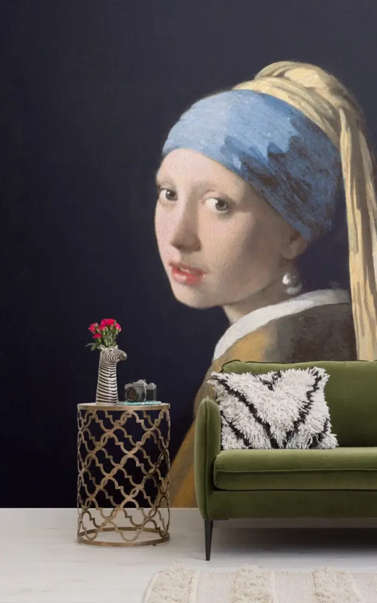 A stylish living area with a dark blue wall featuring a large replica of Vermeer's "Girl with a Pearl Earring," complemented by a vibrant green velvet sofa and an ornate side table displaying a whimsical zebra vase with flowers, enhancing the blend of classical and modern decor elements.