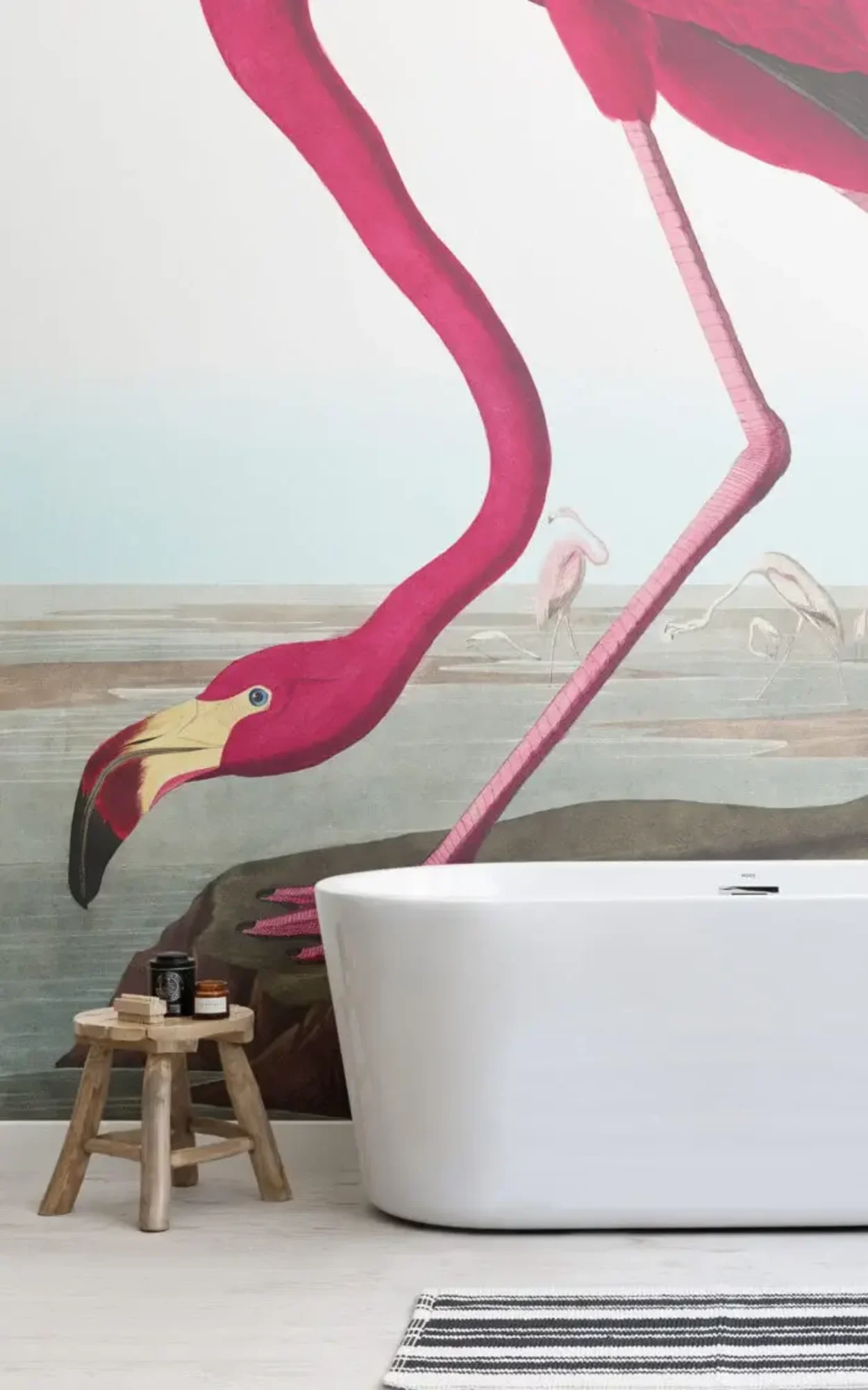 A striking modern bathroom featuring a sleek white freestanding bathtub, a rustic wooden stool with toiletries, and a dramatic mural depicting an oversized pink flamingo, creating a bold and vibrant visual statement in the space.