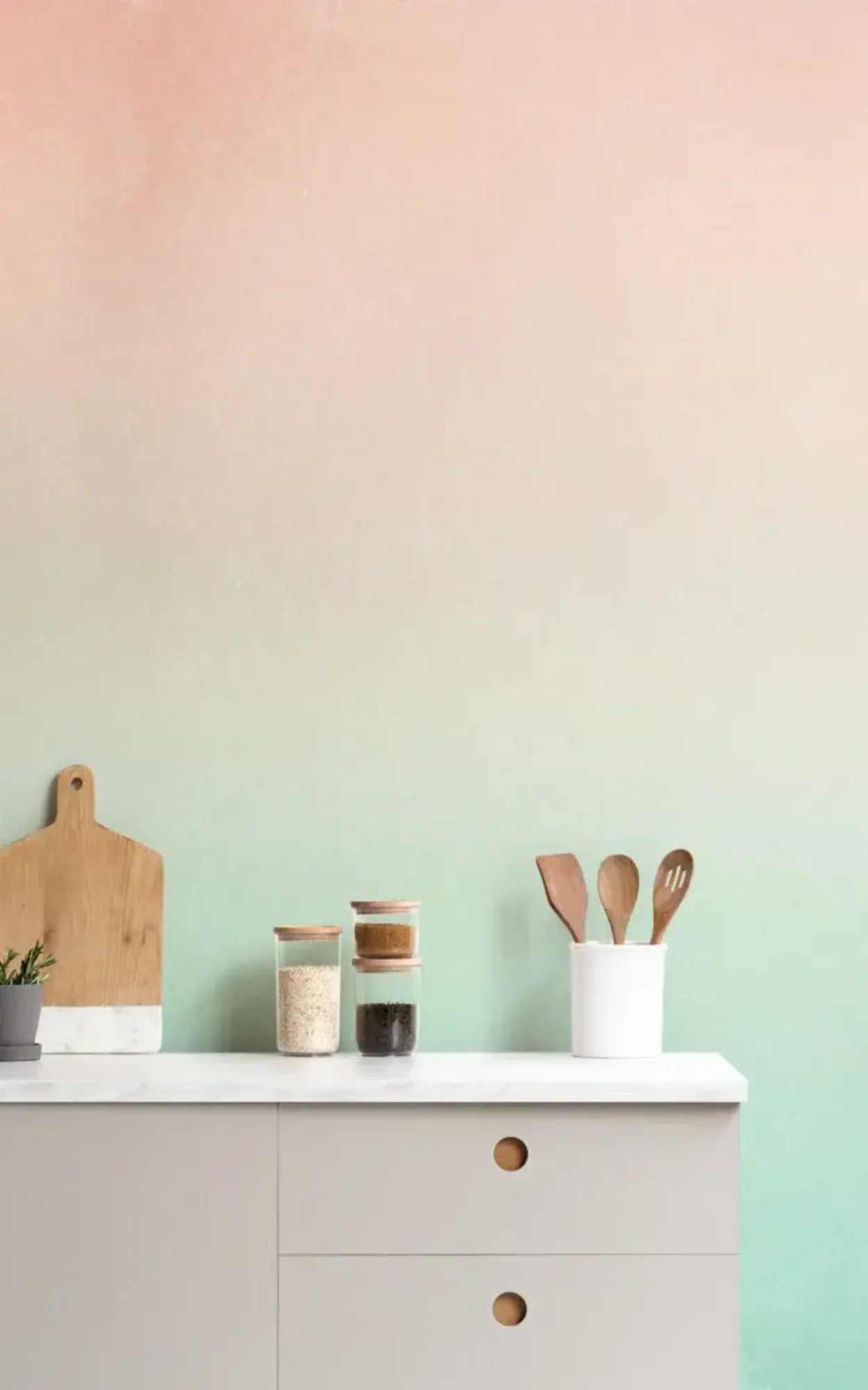 A minimalist kitchen setup featuring a modern white sideboard against a softly blended ombre wall in shades of peach to light green. The sideboard is adorned with a wooden cutting board, three glass jars filled with kitchen staples, and a white container holding wooden cooking utensils, creating a clean and organized aesthetic.