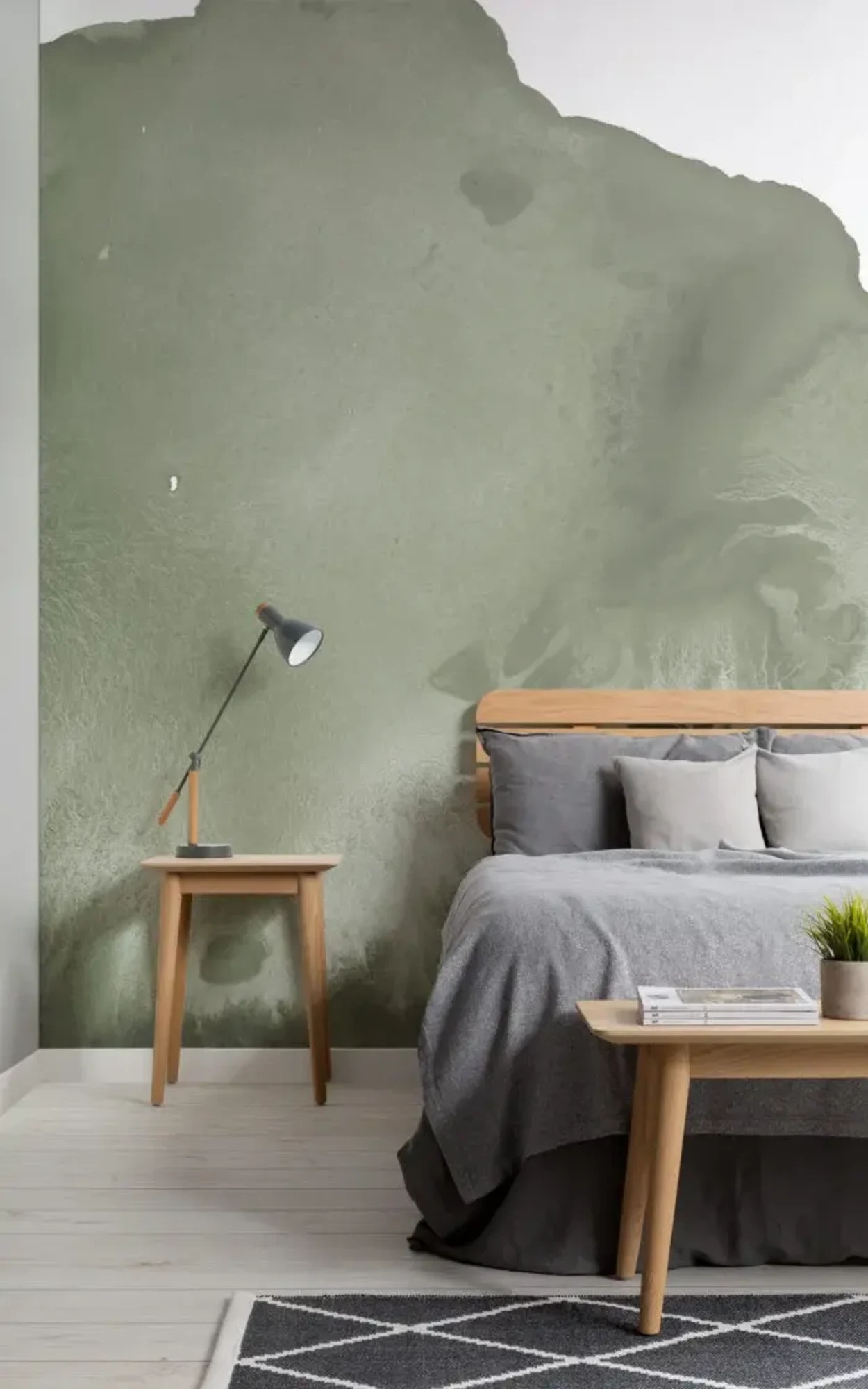  A serene bedroom setting featuring a wooden bed with gray bedding, a small wooden side table with a modern lamp, set against a textured green mural creating a calming and natural backdrop. A black and white patterned rug on the floor completes the peaceful, contemporary aesthetic.
