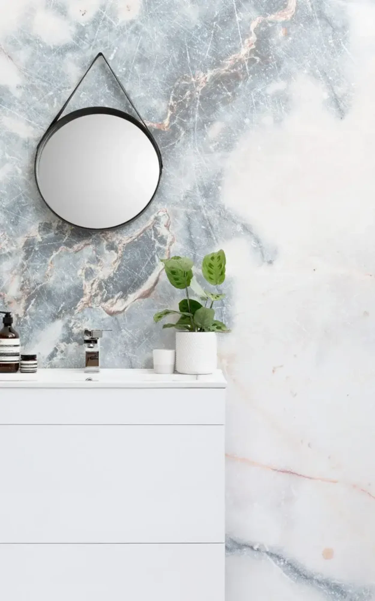 A chic and modern bathroom setup featuring a sleek white vanity against a stunning marble wall in hues of white and blue with pink veining. The scene is elegantly accessorized with a contemporary round mirror, small potted plants, and minimalist decor items, creating a tranquil and sophisticated atmosphere.