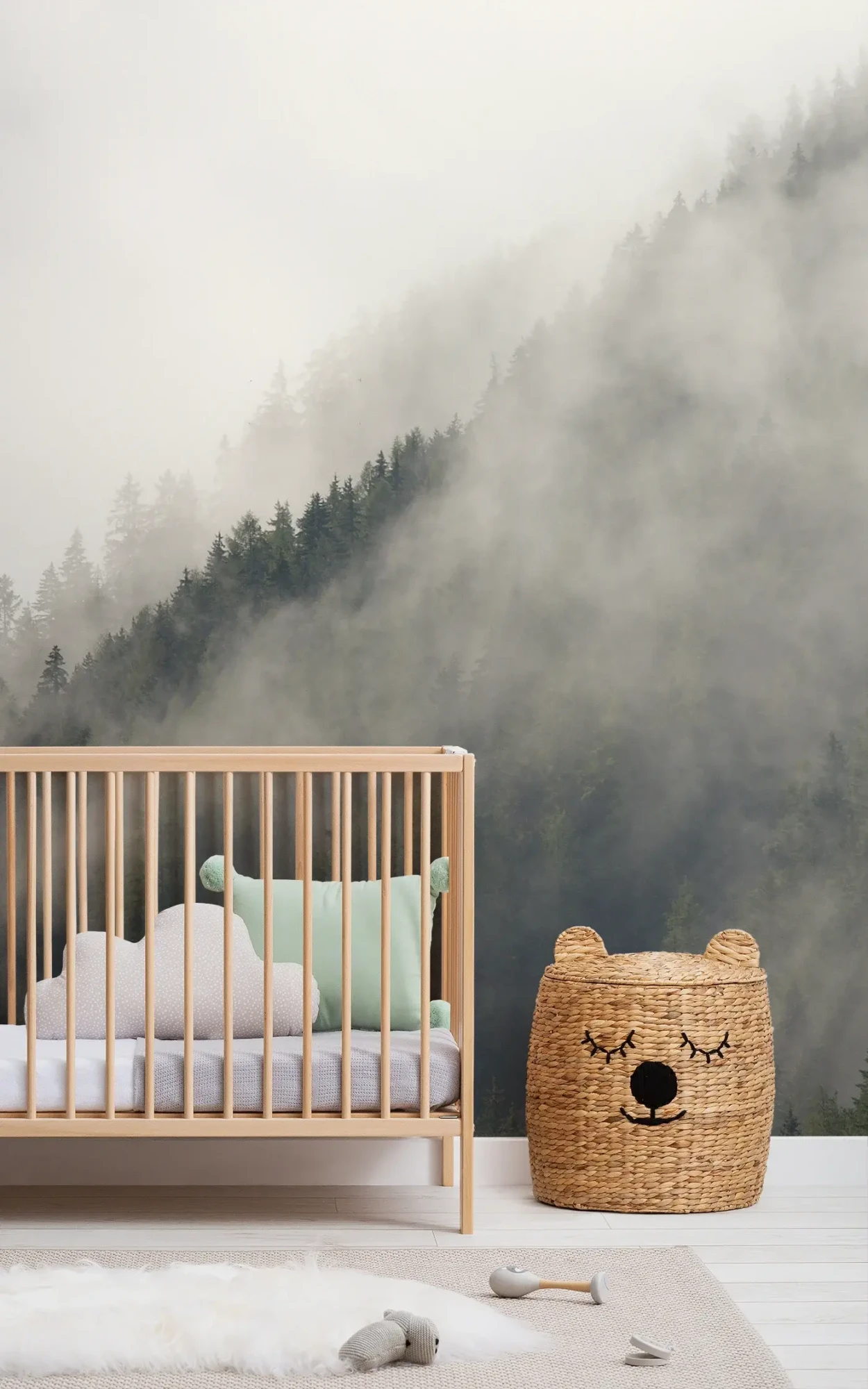 A serene nursery setup with a wooden crib filled with light blue and white bedding, next to a woven bear-shaped storage basket, set against a mural of a foggy, tree-covered mountain landscape.