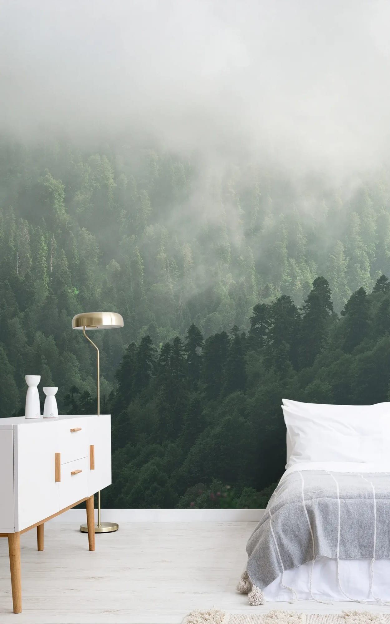 Modern bedroom interior with a white side table featuring a golden lamp and ceramic vases, next to a bed with white bedding and a grey tassel throw, set against a mural of a misty, densely forested mountain landscape.