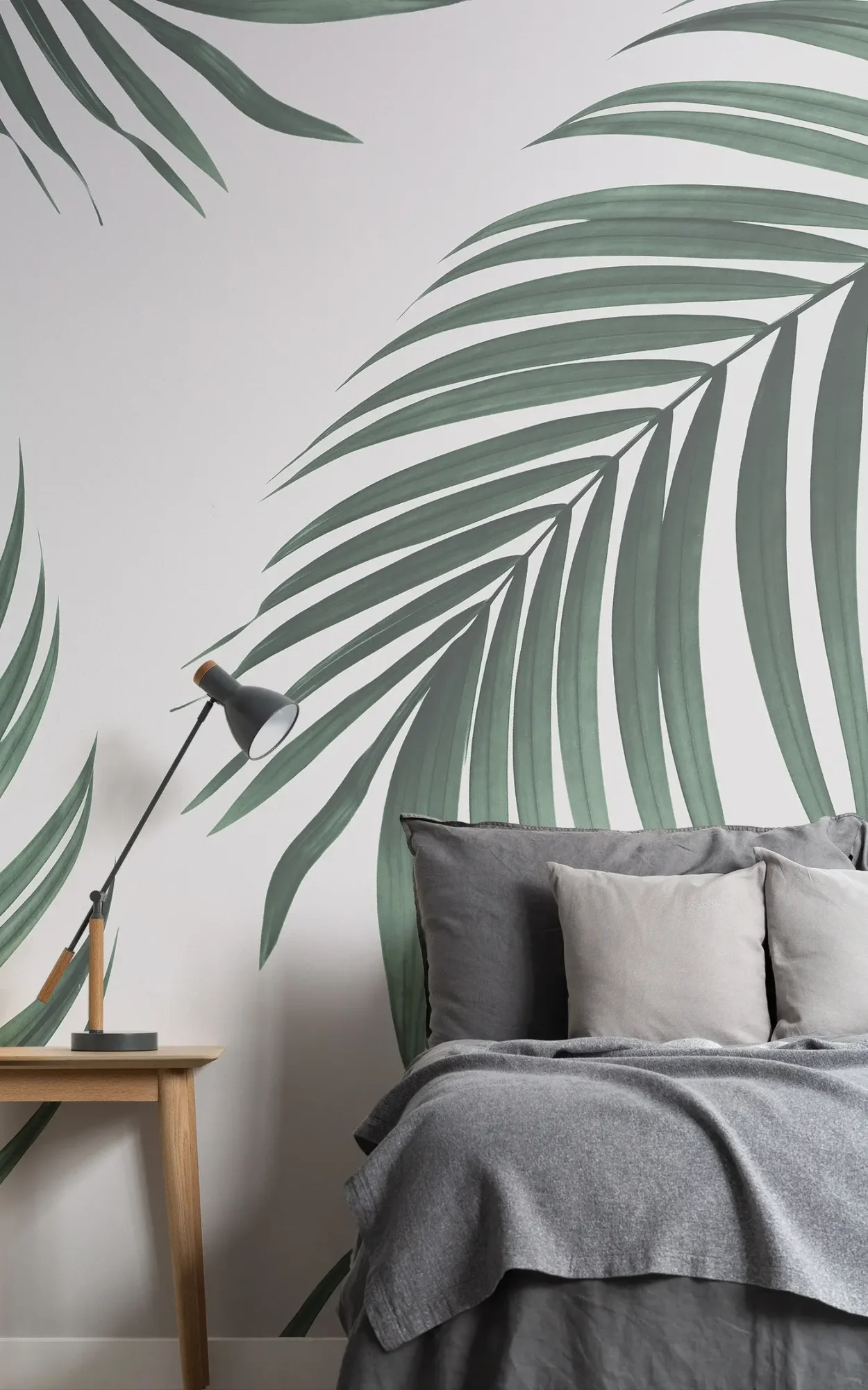 Modern bedroom with a bed dressed in gray linens and multiple pillows, beside a wooden nightstand with a gray lamp, against a wall decorated with large green palm leaf patterns.