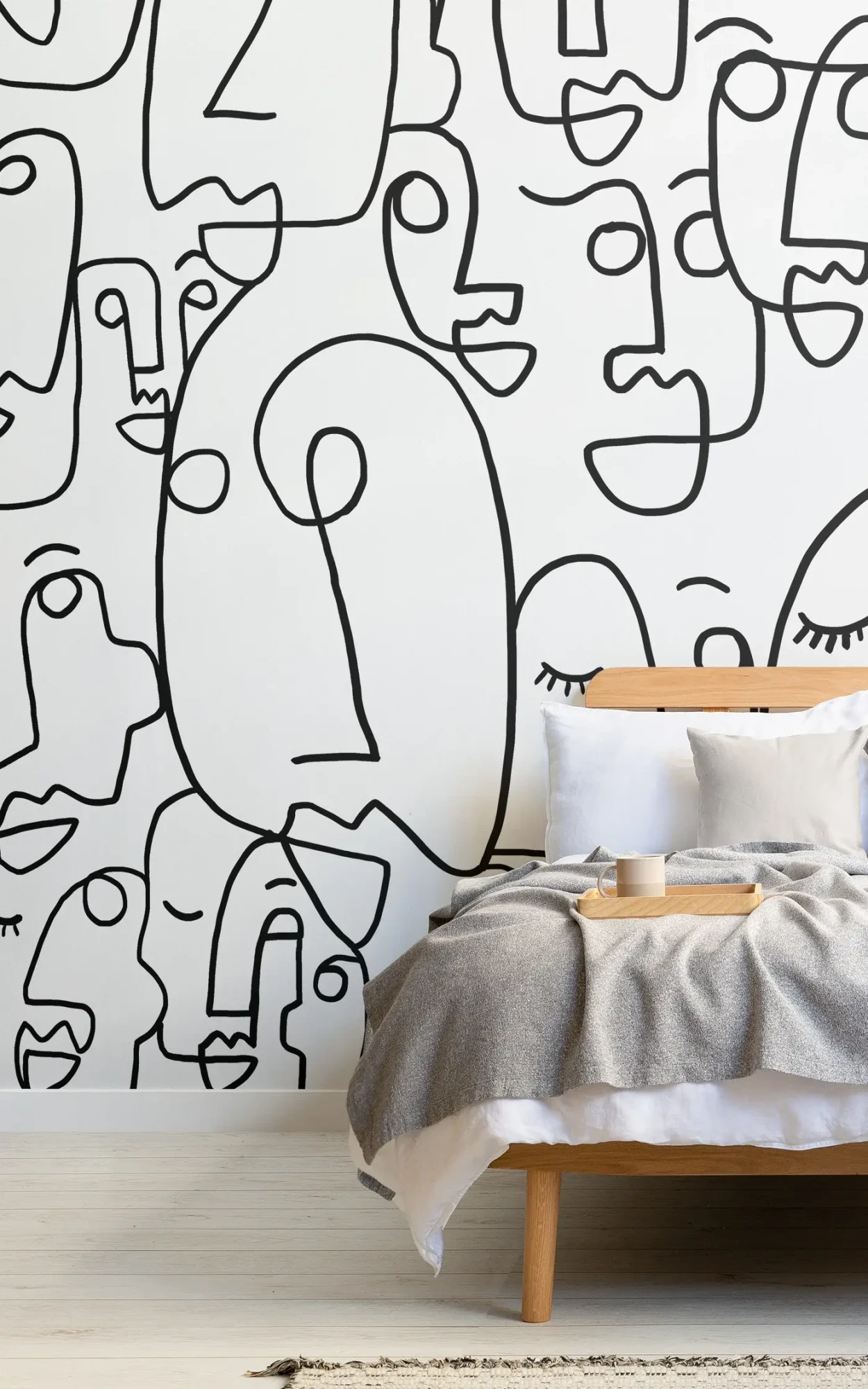 Minimalist bedroom design featuring a bed with a wooden headboard and gray bedding, next to a wooden nightstand with a tray, against a large wall mural of black and white abstract face doodles.