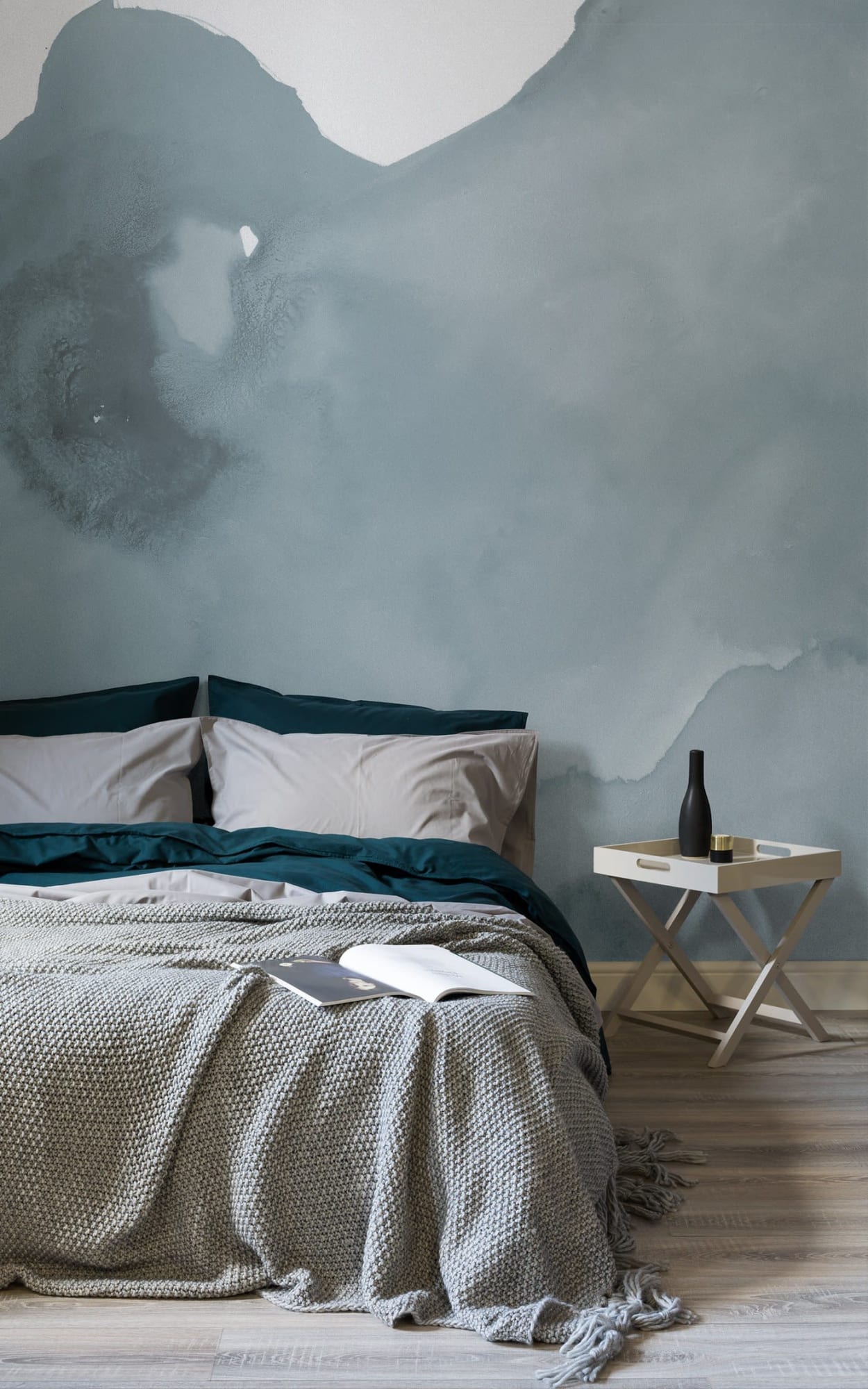Contemporary bedroom featuring a bed with dark green and beige pillows, a chunky grey knitted throw, beside a white side table with a dark vase, against a textured grey wall.