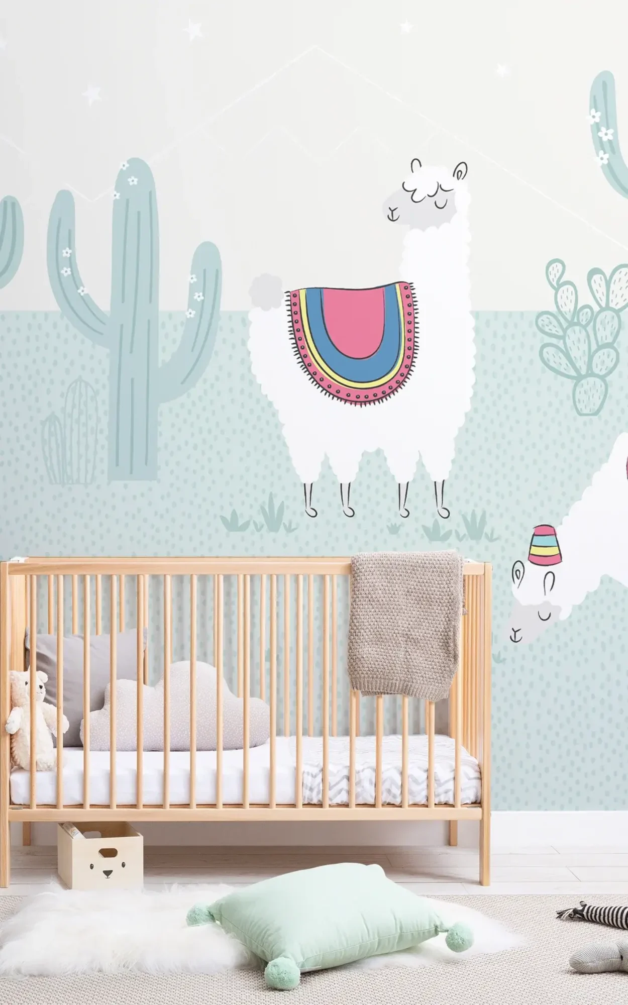 A nursery featuring a wooden crib beside a knitted throw, with a whimsical wall decorated with cartoon cacti and llamas against a dotted pale green background.