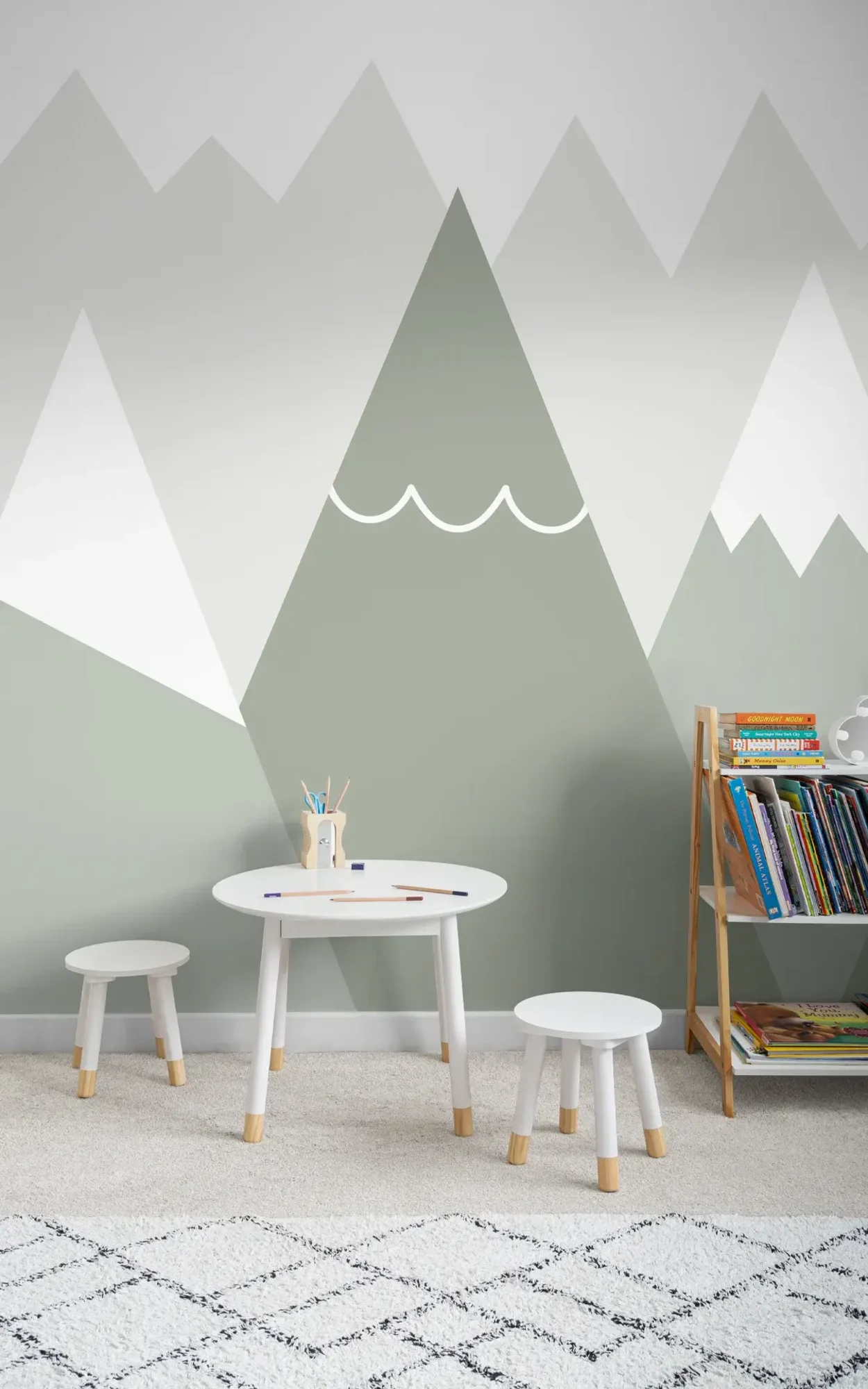 A children's play area with a small white table, two stools, and a bookshelf against a wall painted with a geometric mountain pattern in shades of white and green.