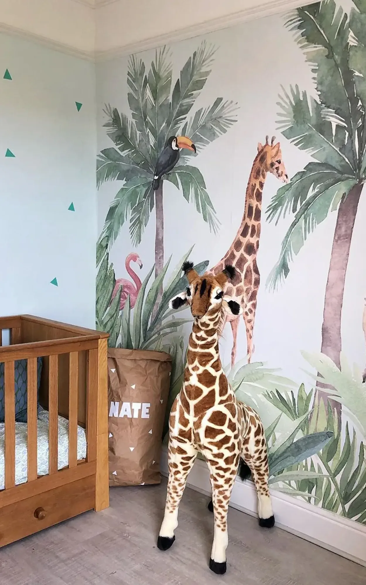 Children's nursery featuring a wooden crib, a large stuffed giraffe, and a colorful mural of a tropical scene with palm trees, a toucan, and a giraffe.