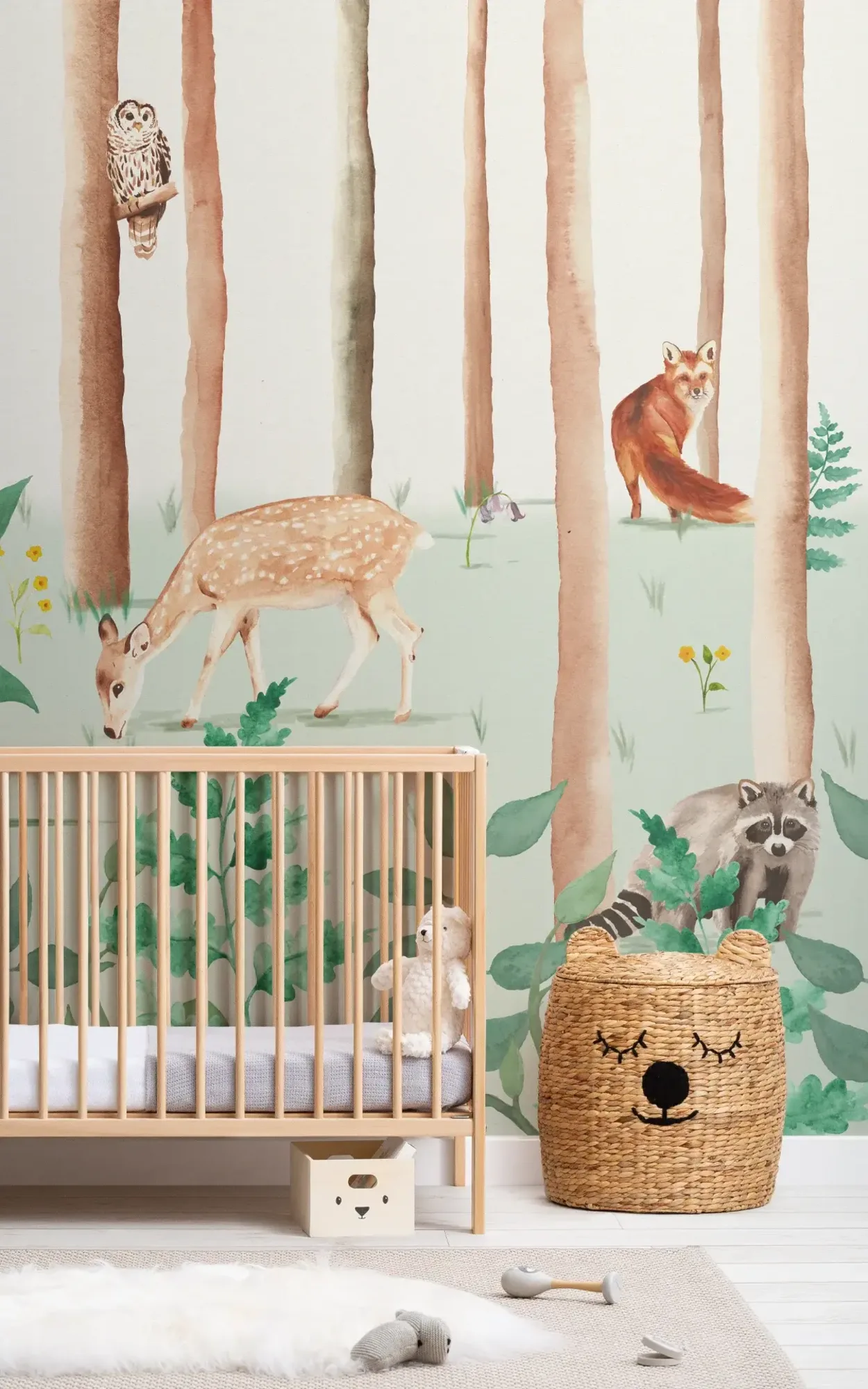 Charming nursery room featuring a wooden crib against a whimsical wall mural of a forest scene with trees, a deer, fox, raccoon, and owl, accompanied by a woven bear-shaped storage basket and plush toys on the floor.