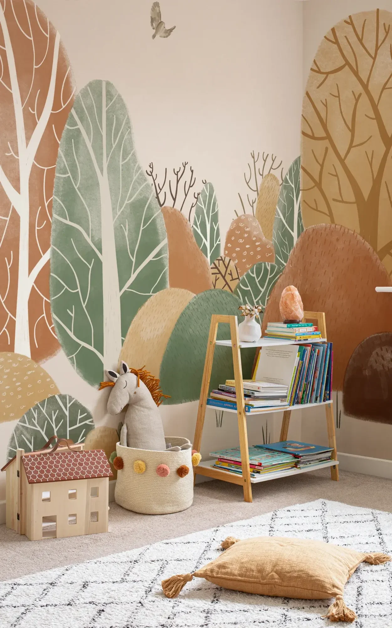 Playful children's reading nook with a bookshelf filled with colorful books, a soft toy unicorn in a woven basket, and a child-sized wooden house, against a whimsical mural of abstract, colorful hills and trees.