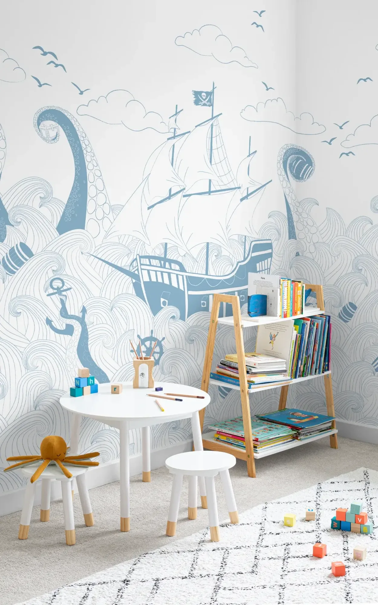  A children's play area with a small white table and stools, a bookshelf filled with colorful books, set against a large mural depicting a whimsical ocean scene with a pirate ship and sea monster.