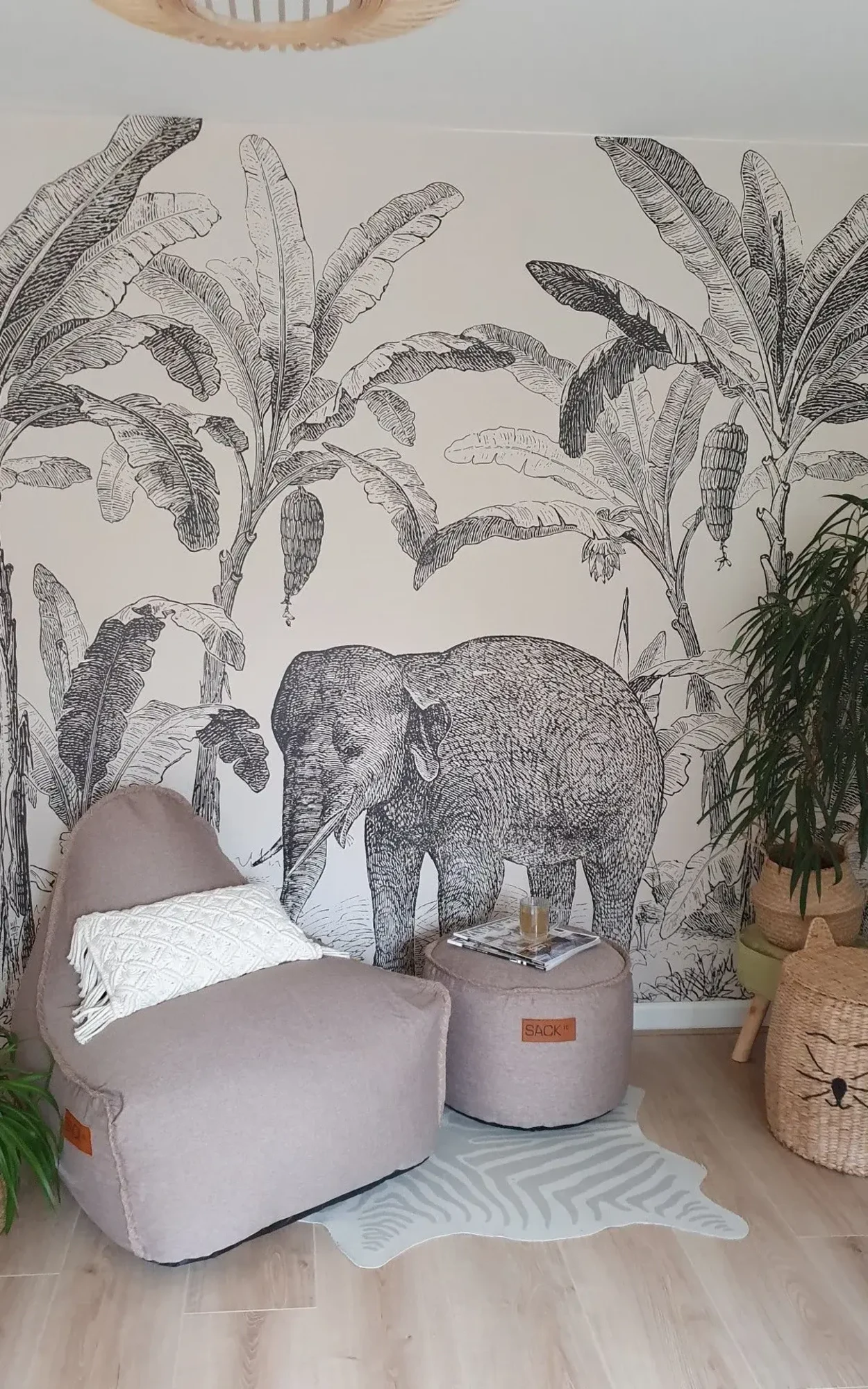 A cozy reading nook featuring a plush chair and ottoman in beige, a small round table with a drink, set against a detailed wall mural depicting an elephant and lush tropical foliage in black and white.