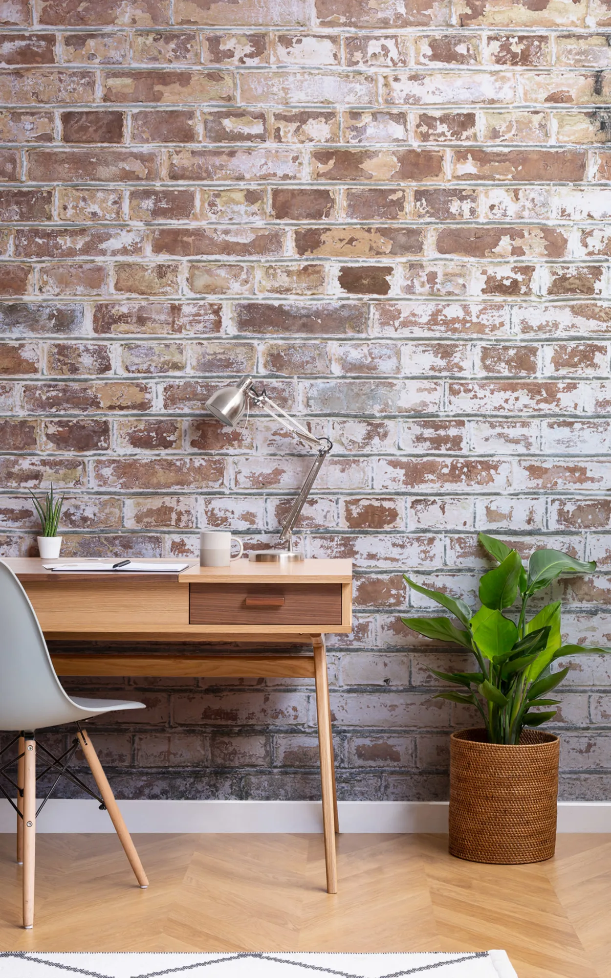 Home office with weathered brick wall mural, wooden desk, metal lamp, and green potted plant.