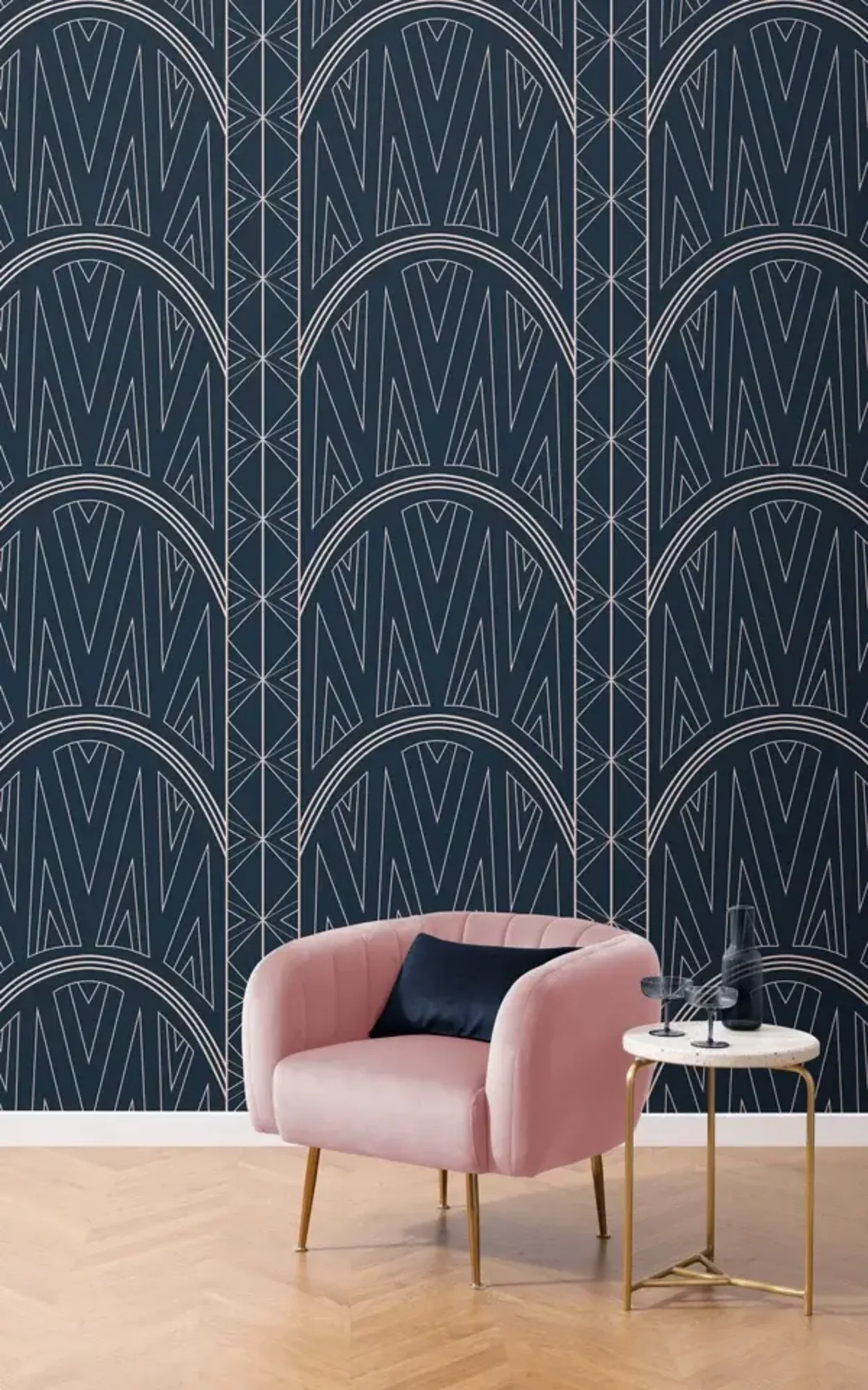  A stylish interior featuring a plush pink armchair with a dark blue cushion beside a small round gold-trimmed table, set against a backdrop of dark blue wallpaper with a large geometric Art Deco design in white lines.